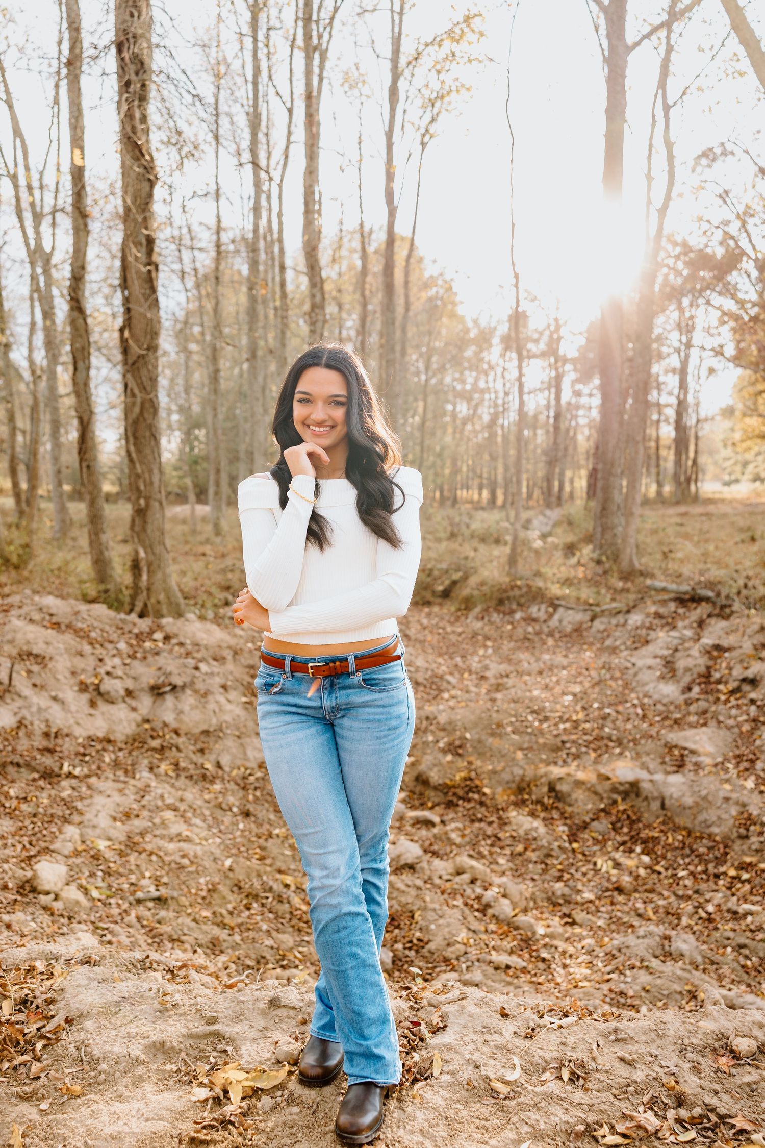 A person in a white sweater and jeans poses in a sunlit autumn forest with fallen leaves on the ground.