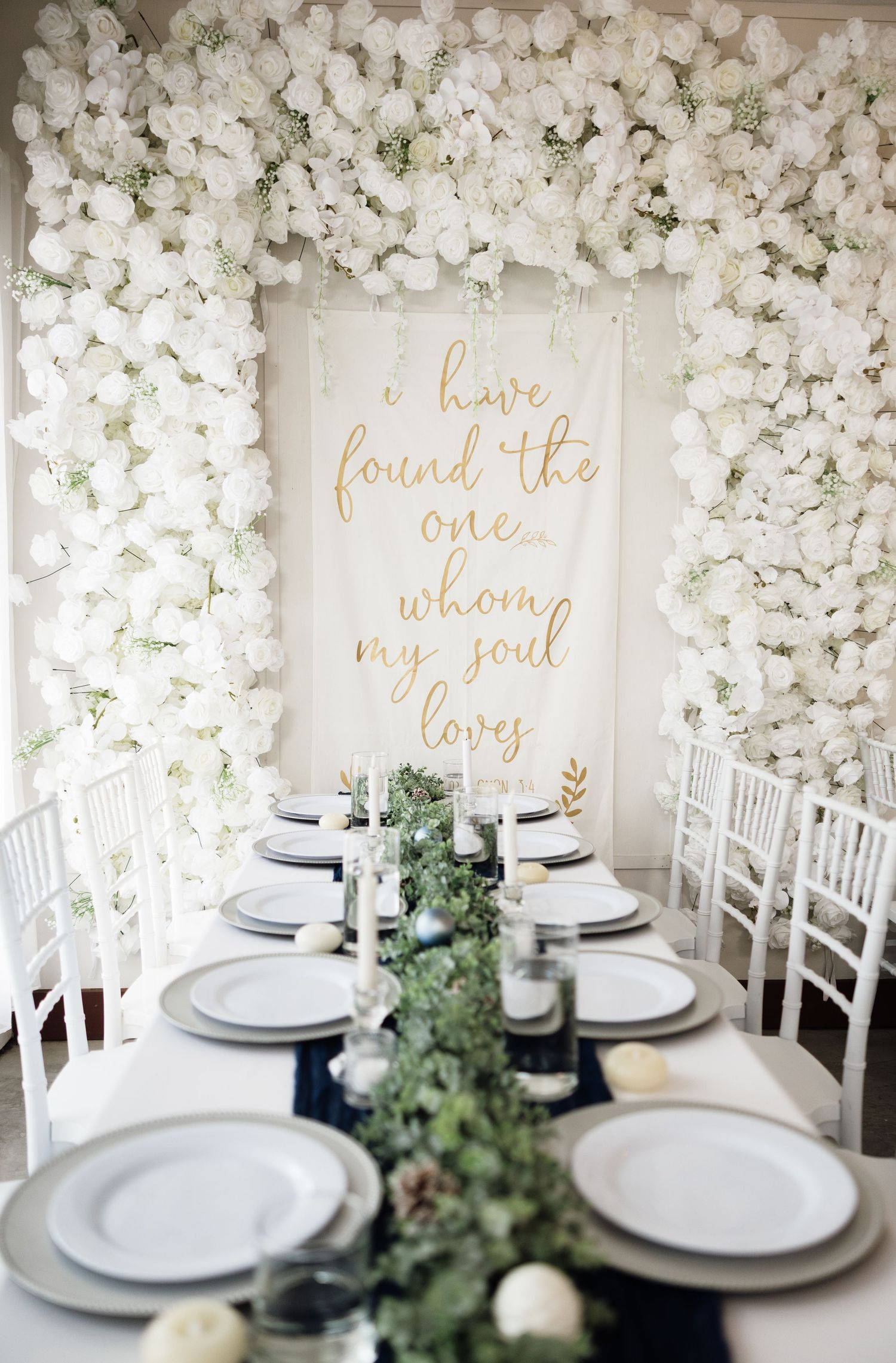 Elegant wedding reception table setup with white floral wall backdrop and romantic gold text decor.