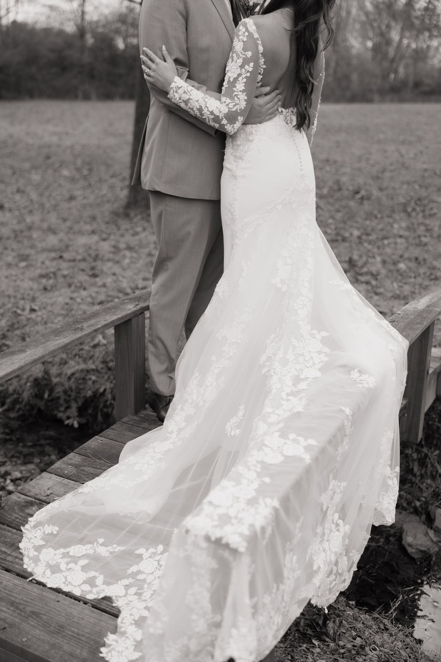 Wedding couple embracing on a wooden deck with a detailed lace dress trailing behind.