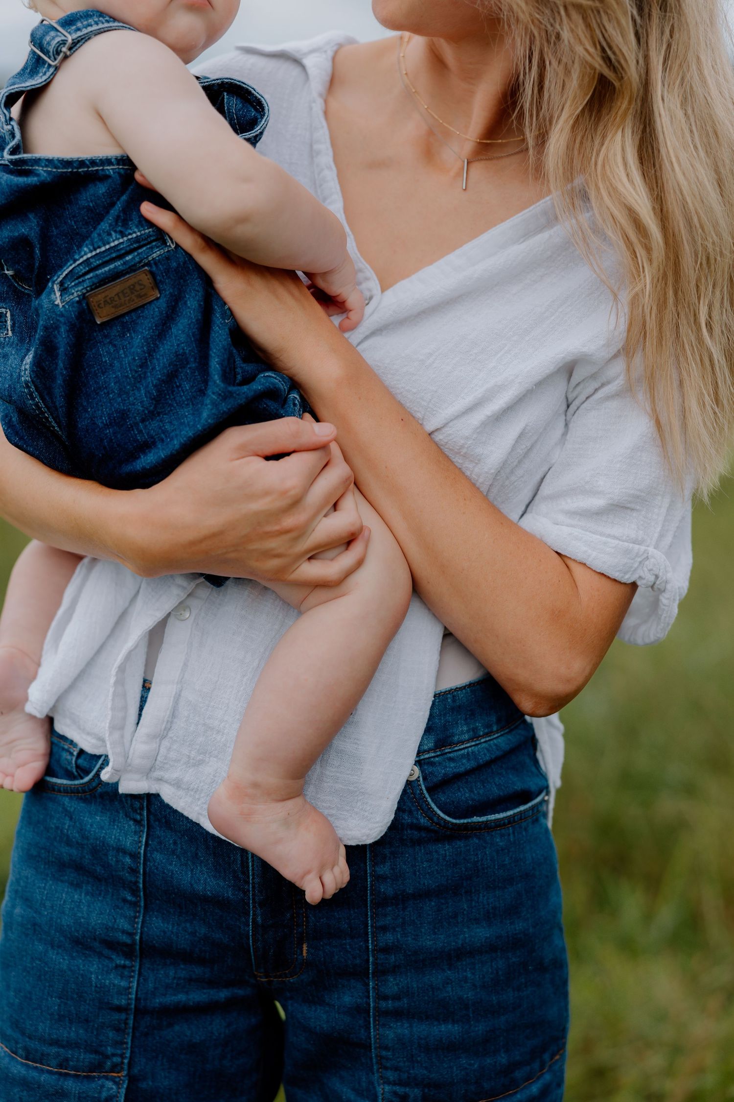 Close-up of parent in white shirt holding small child wearing denim overalls outdoors in summer setting.