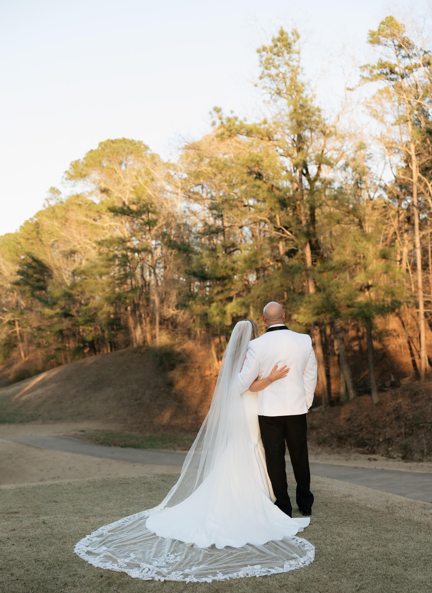 Wedding couple photographed from behind with flowing lace veil against natural outdoor backdrop.