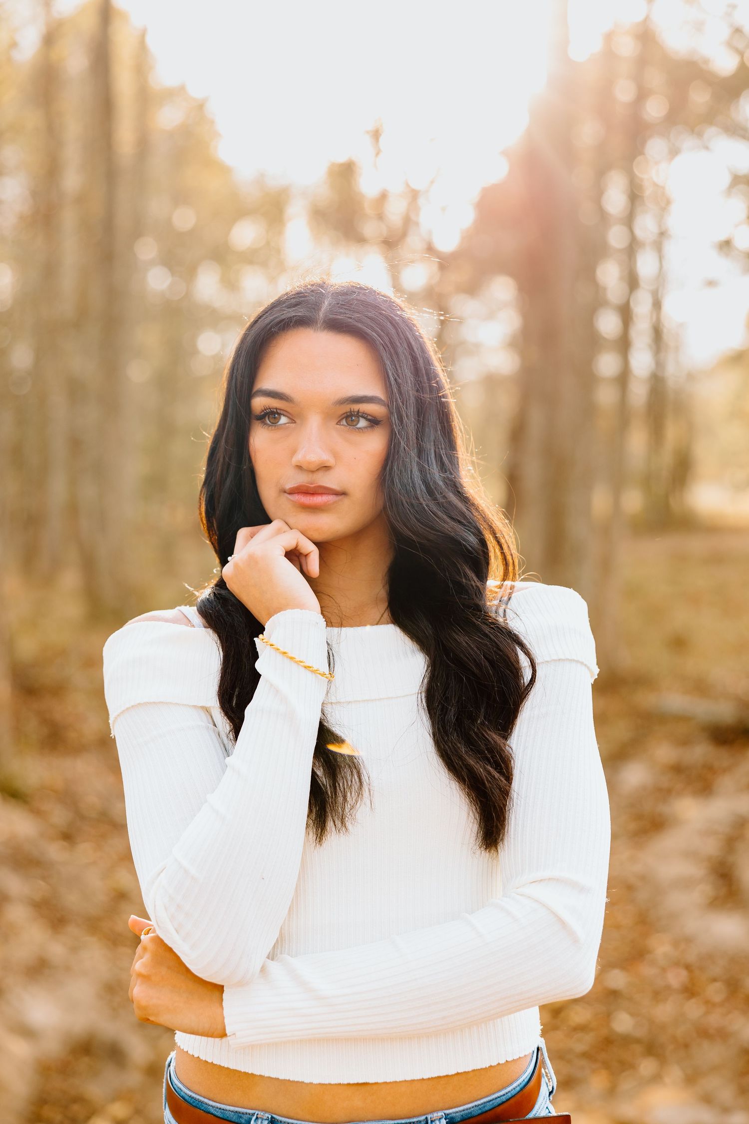 Woman in white sweater poses thoughtfully in autumn forest setting during golden hour sunset.