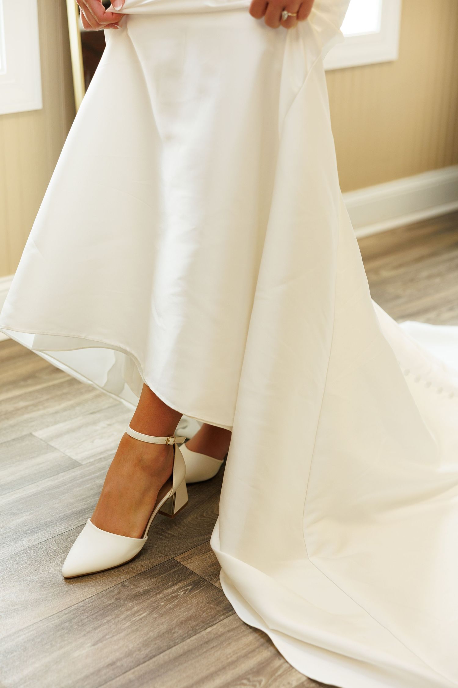 Close-up of elegant white bridal shoes with ankle straps peeking beneath a flowing wedding dress hem on hardwood floor.