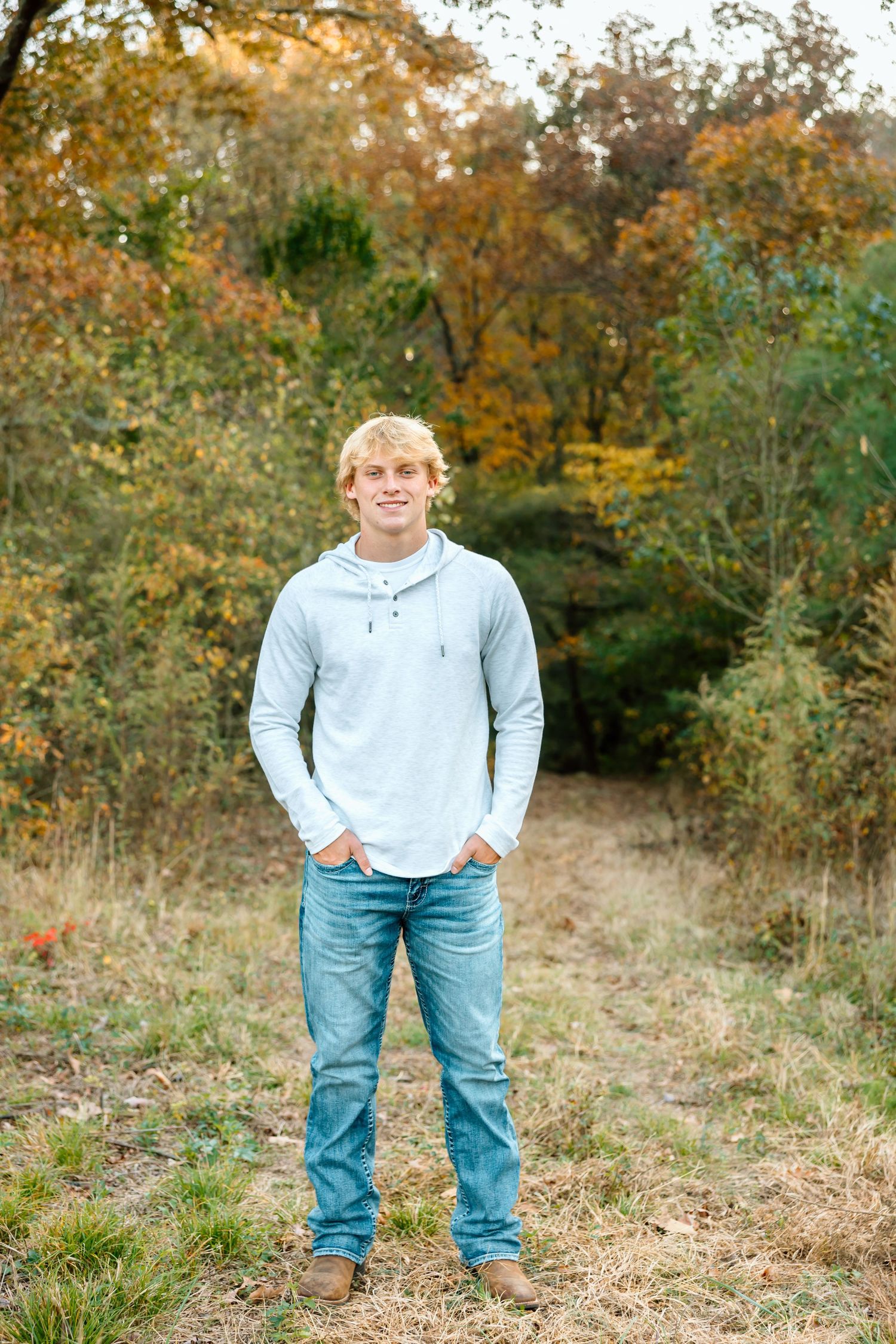 Person in light gray hoodie and jeans standing outdoors in autumn setting with fall foliage in background.