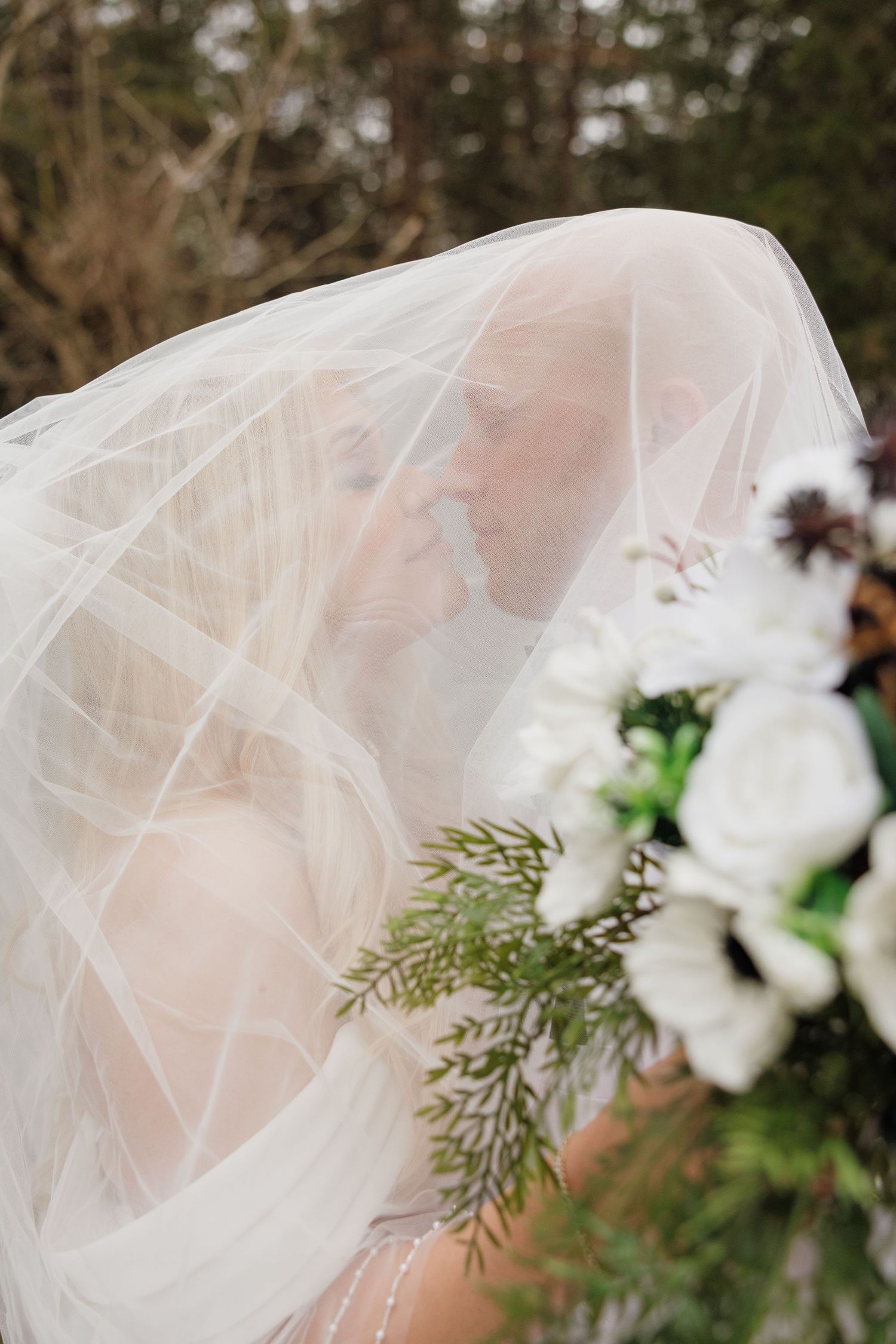 A delicate white wedding veil drapes over a bride's profile with an elegant white floral and evergreen bouquet in view.