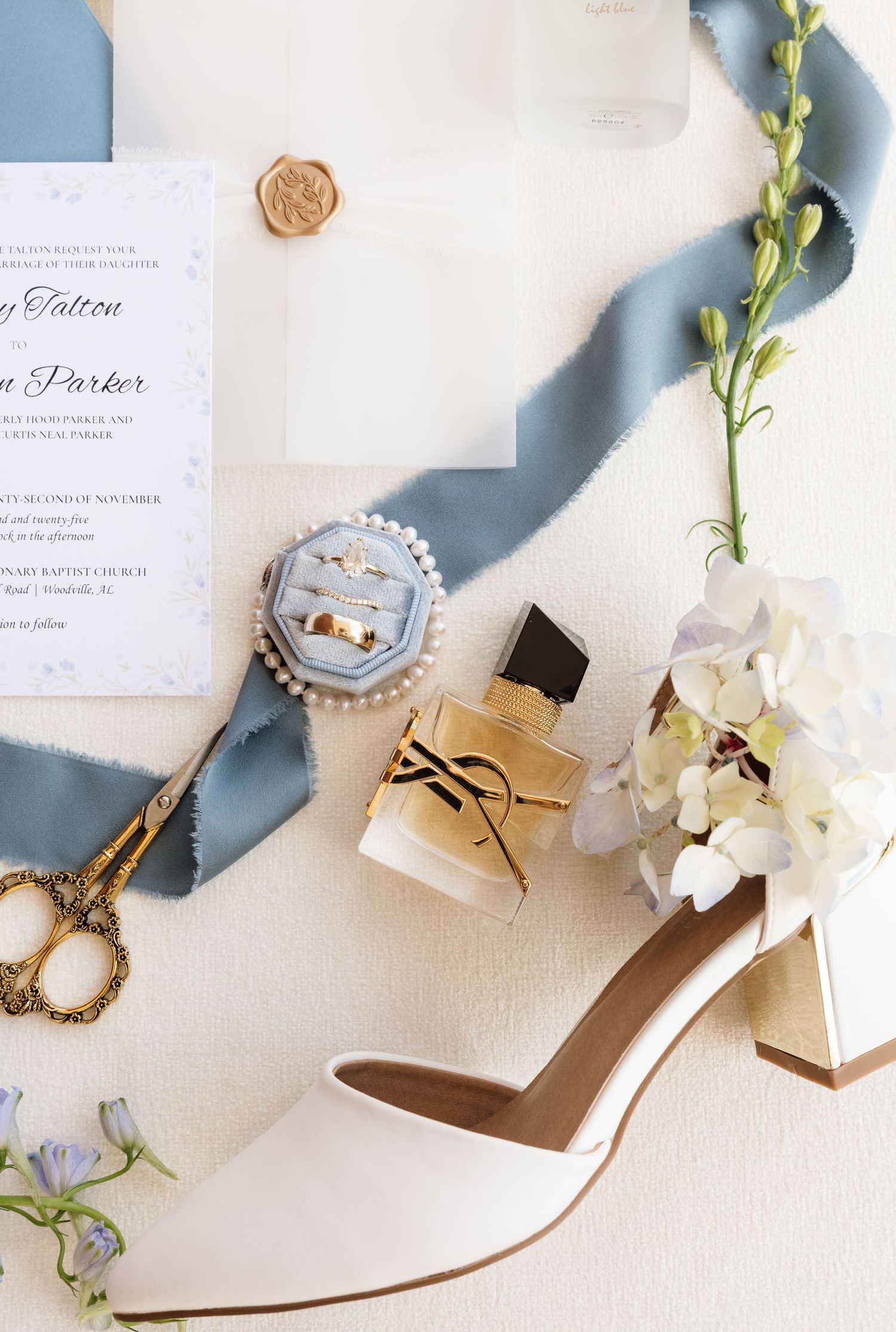 Elegant wedding details including white satin heels, delicate floral stems, and paper invitations arranged on cream background.