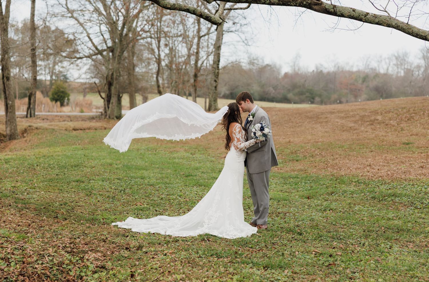 Wedding couple embracing in a field with bride's veil flowing in the wind.