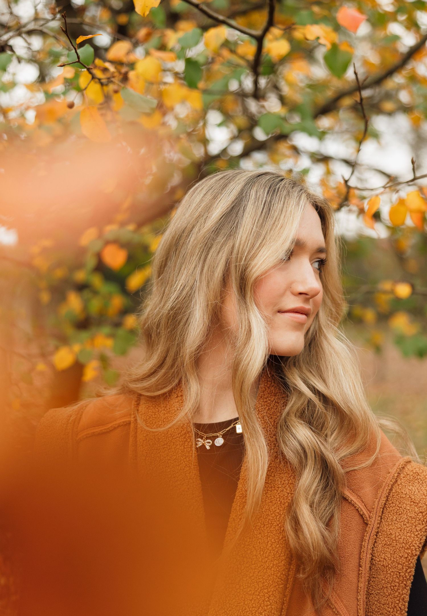 A person with wavy blonde hair poses outdoors against autumn foliage in a rust-colored sweater.