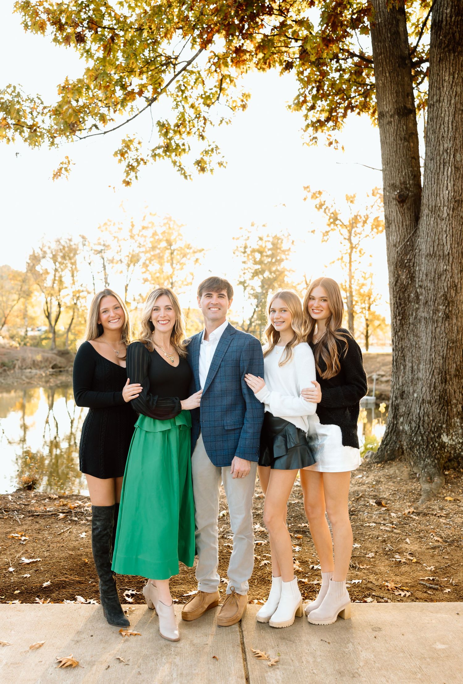 A group stands together under autumn trees by a lake during golden hour wearing stylish outfits in black, green and blue.