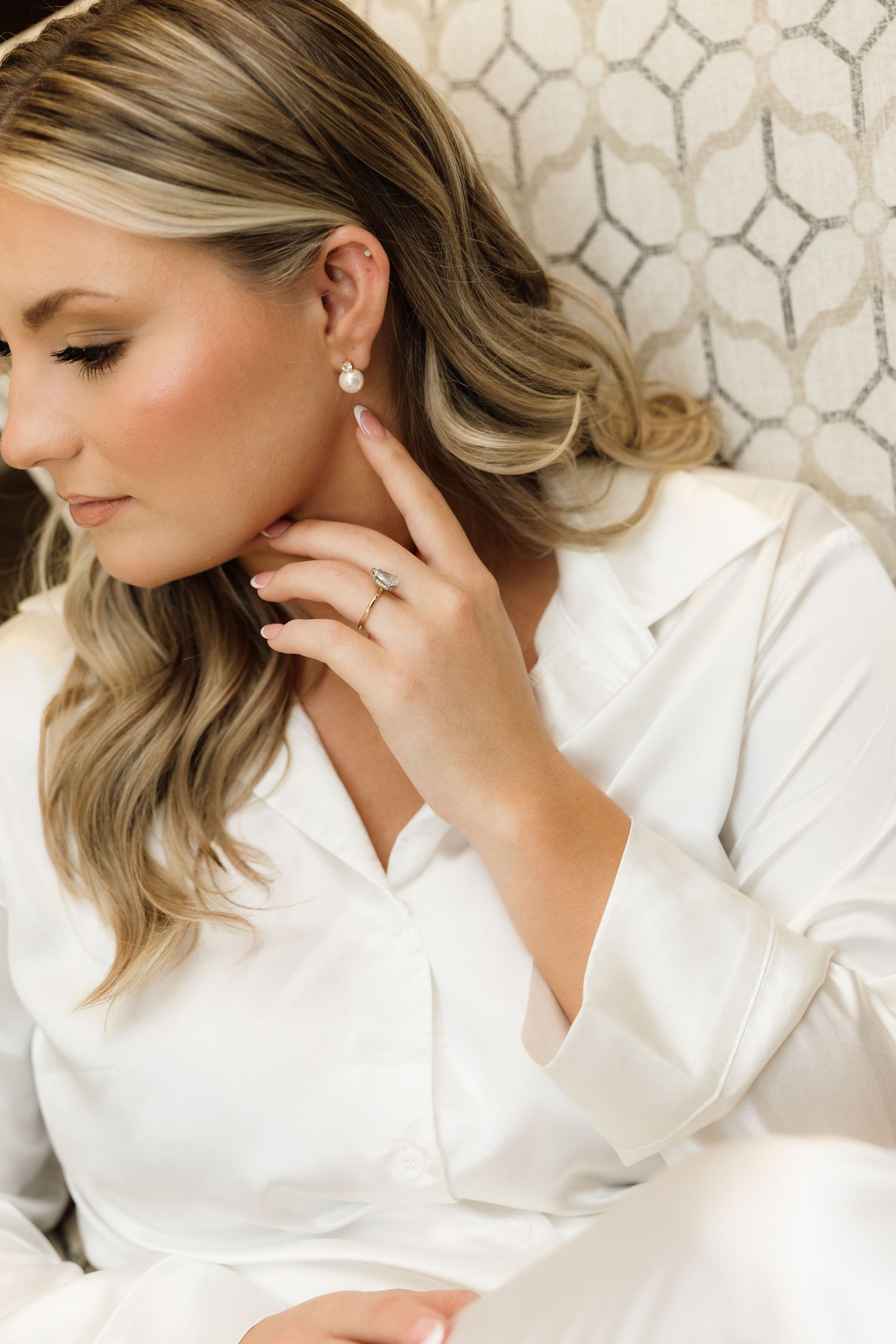 A woman in a white button-down shirt poses elegantly with soft makeup and blonde hair against a patterned background.