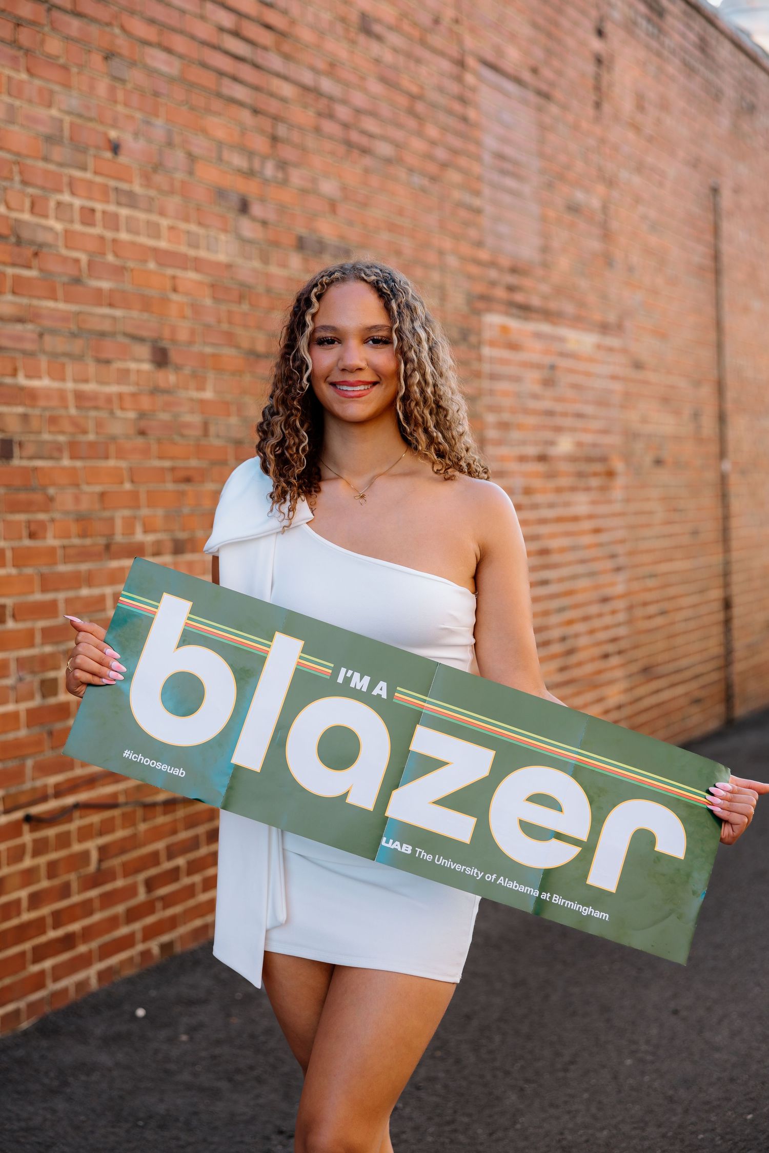 Person in white dress holding green Blazer sign against brick wall.