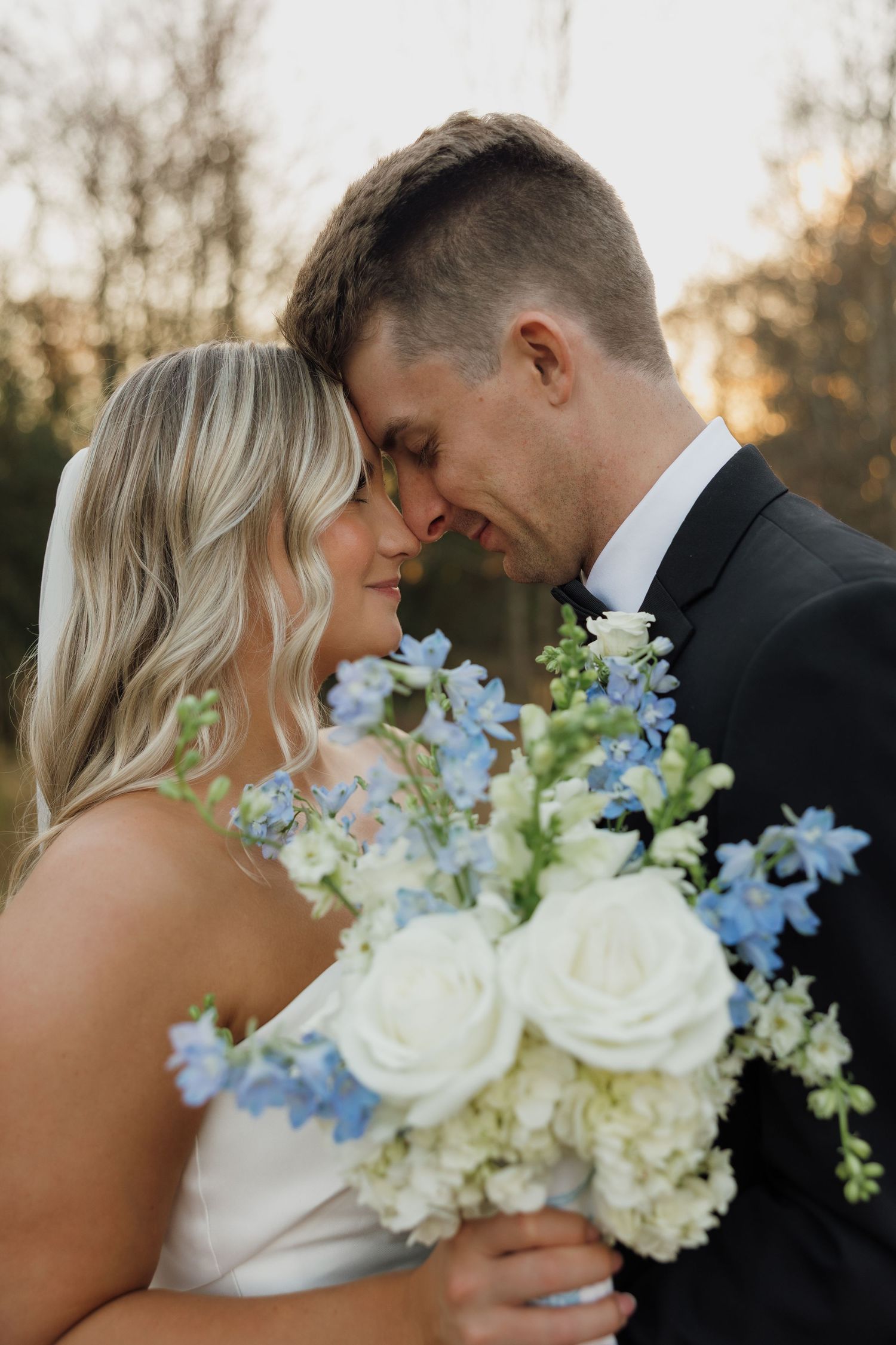 A romantic wedding couple shares an intimate moment at sunset with a beautiful blue and white floral bouquet.