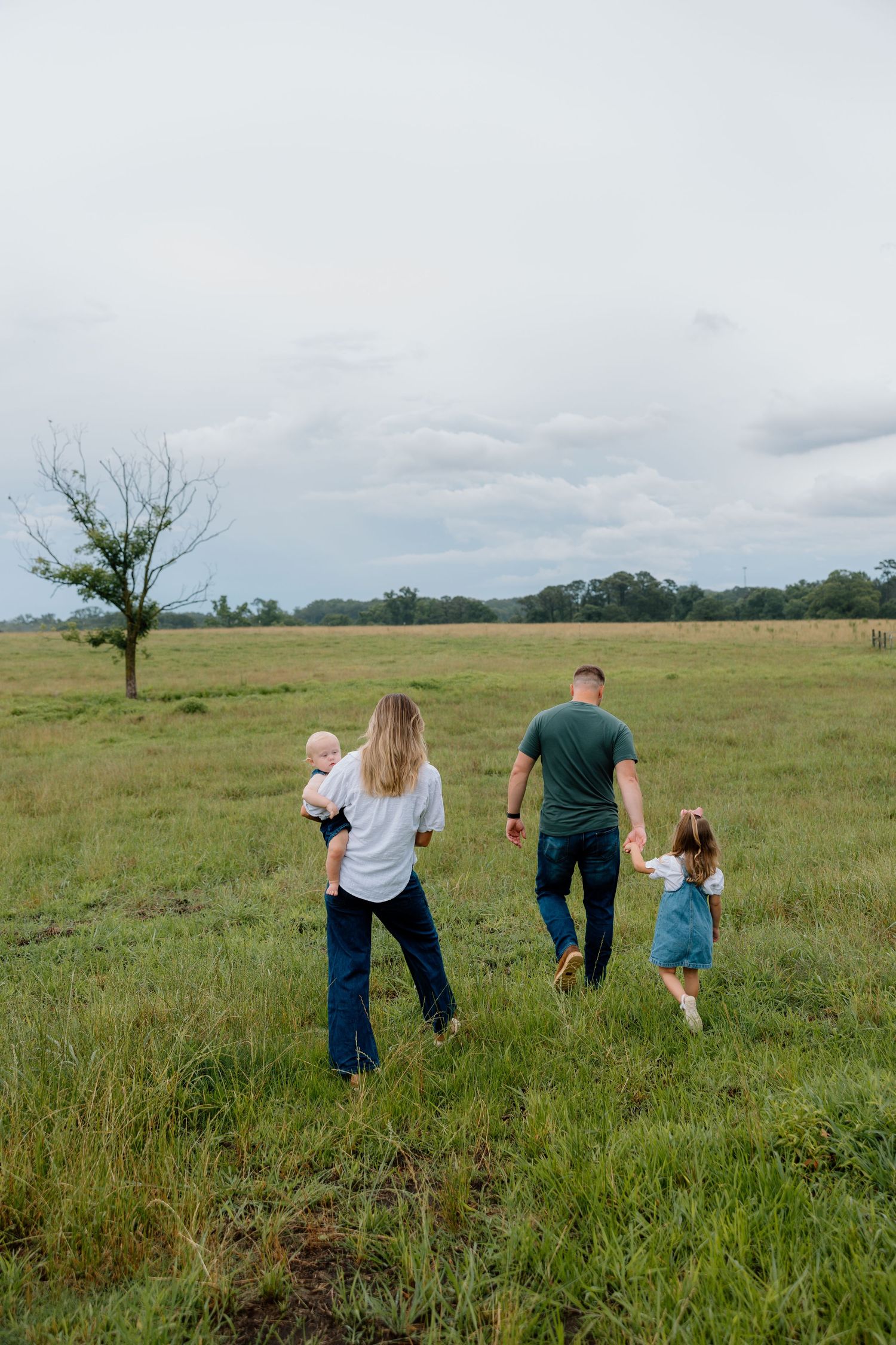 A family walks together through a green meadow with a lone tree and cloudy sky in the background.