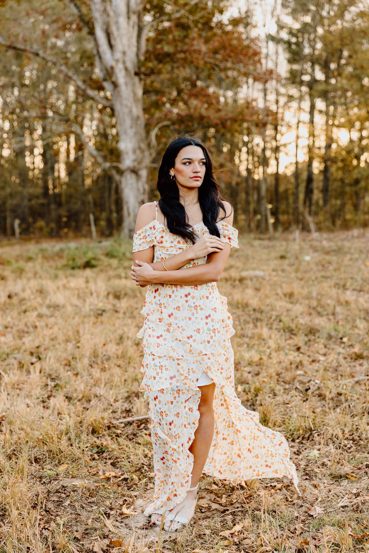 A person in a floral off-shoulder dress poses gracefully in a sunlit field with trees in the background.