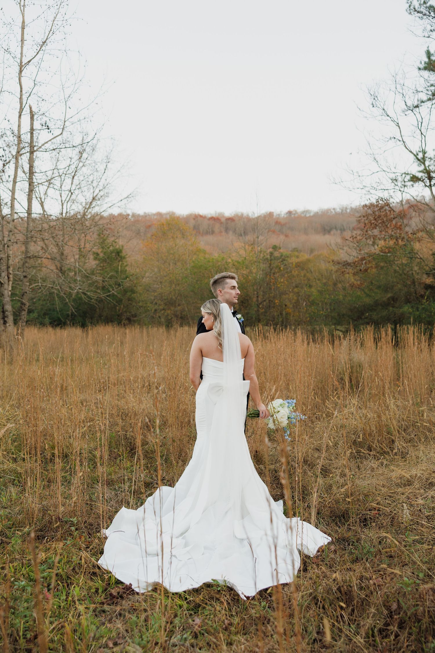 A white wedding dress billows in a rustic autumn field surrounded by tall grass and bare trees.