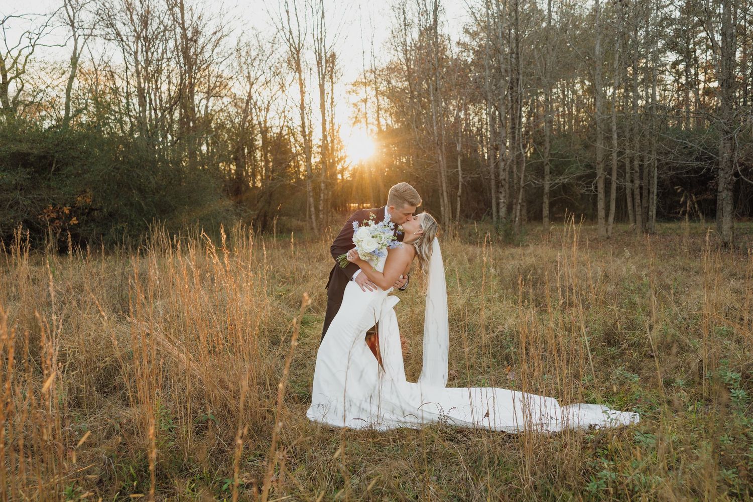 A romantic sunset wedding photo in a golden field with tall grass and bare trees in the background.