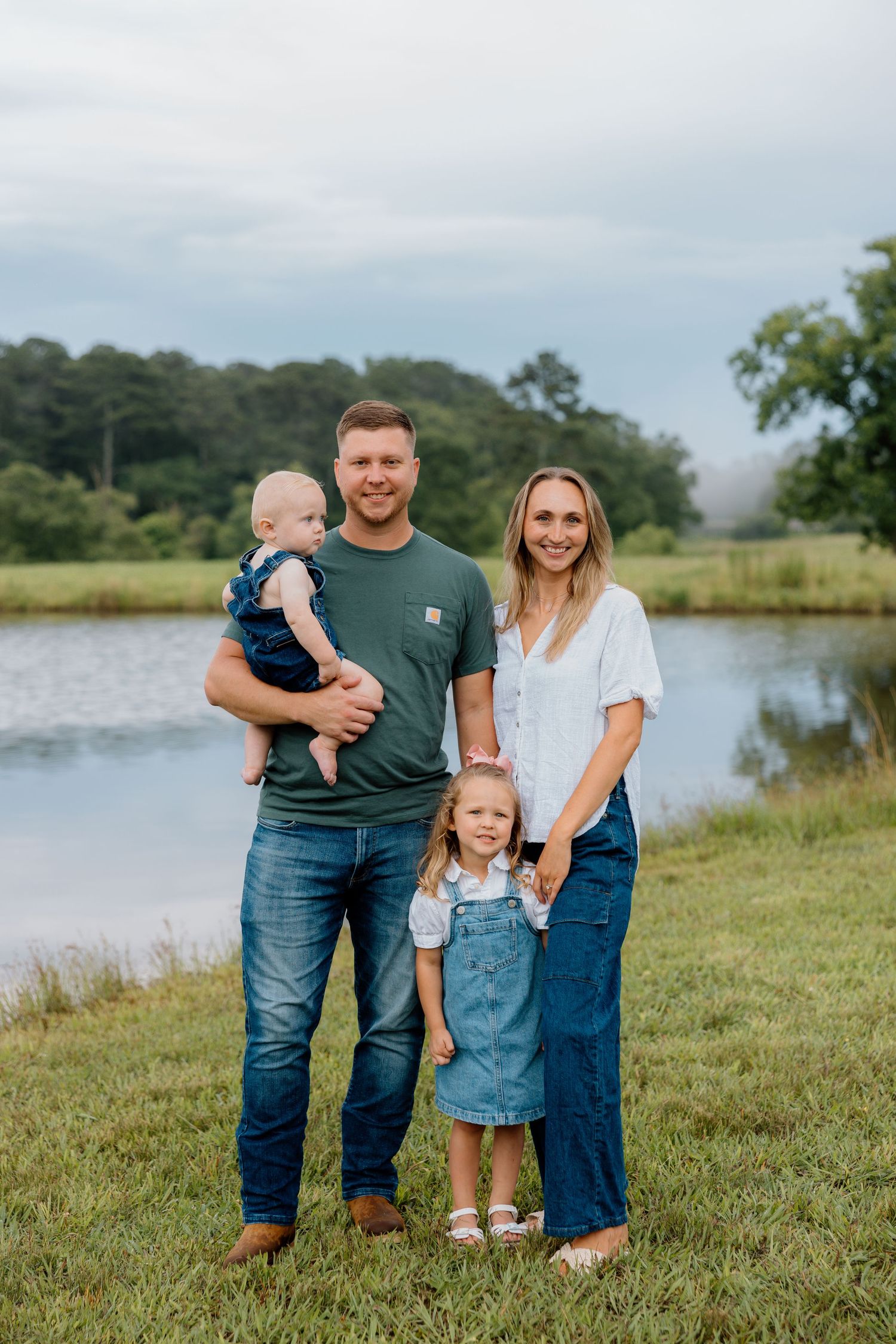 A family stands together by a serene lake on a grassy shore during a peaceful summer afternoon.