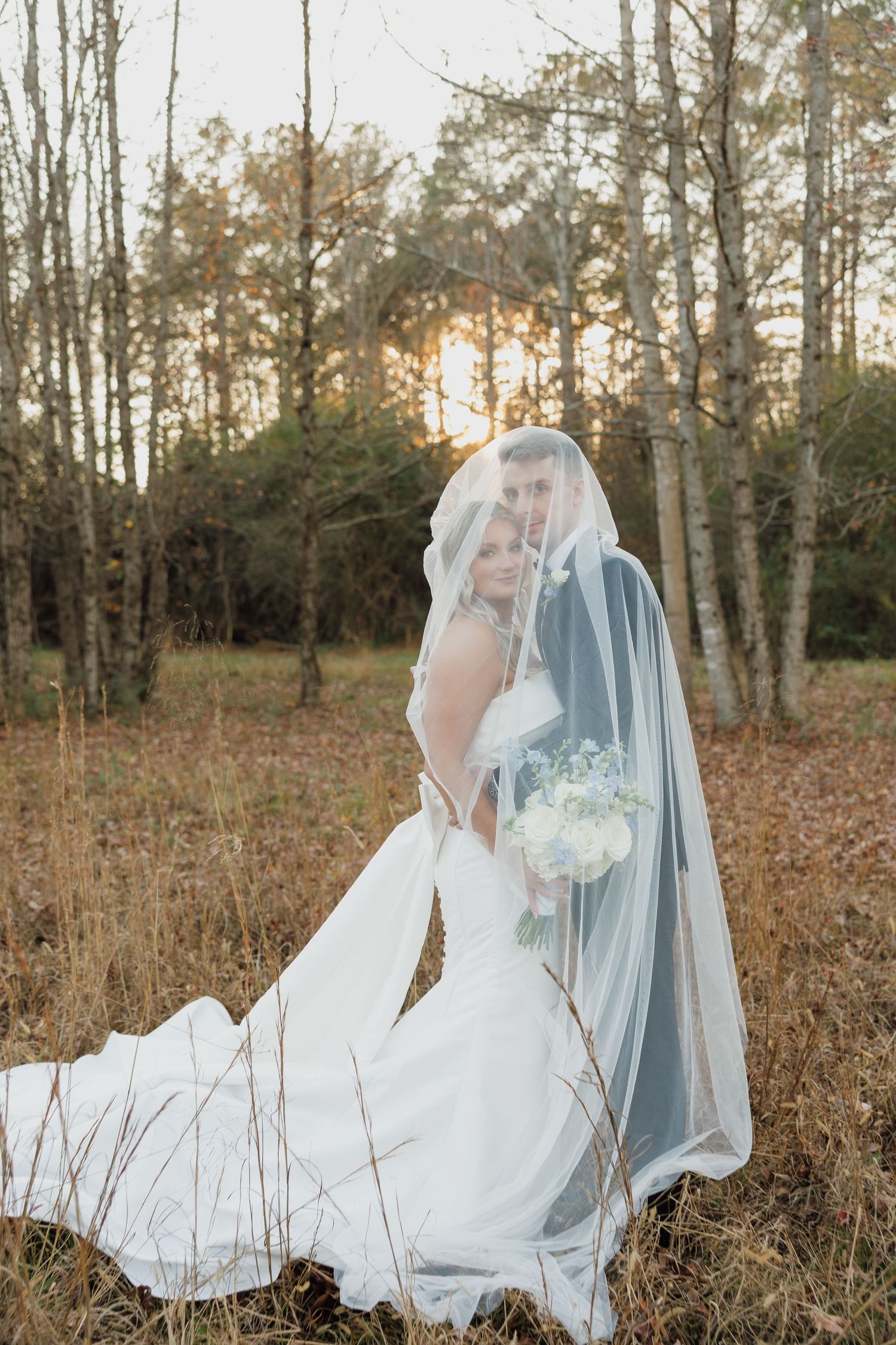 A bride in a flowing white gown poses in a rustic autumn field with fallen leaves.