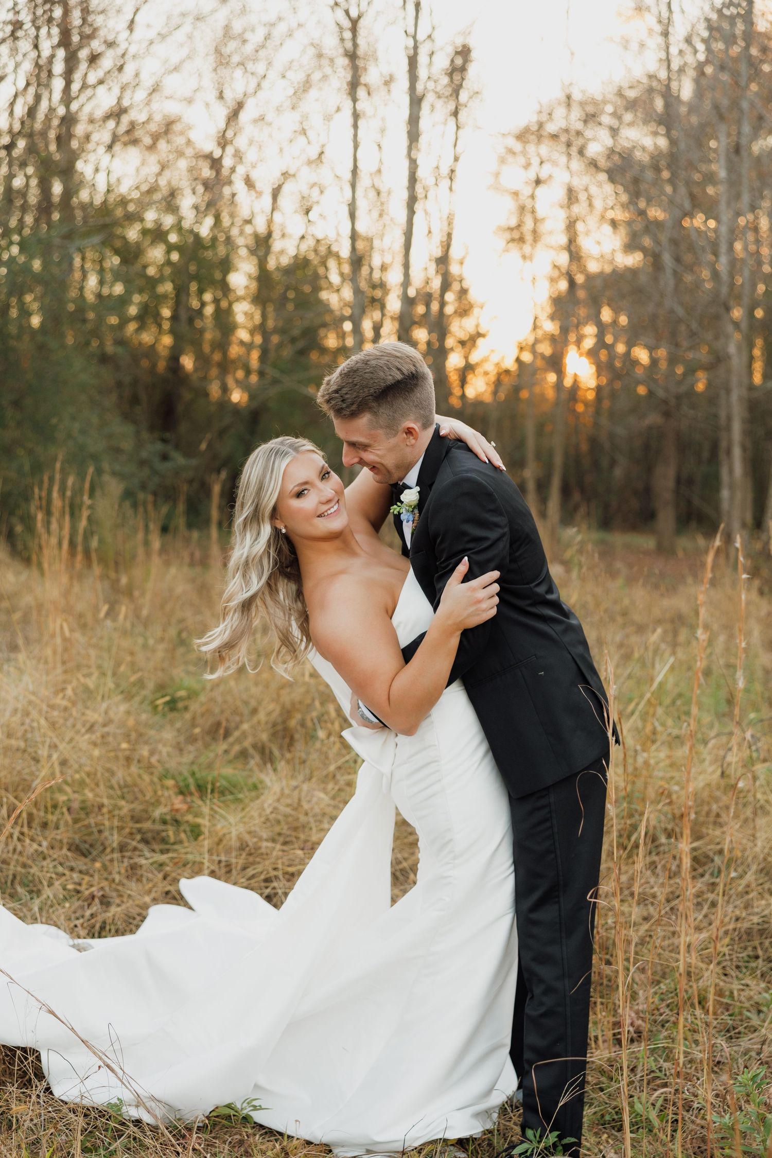 Couple in formal wedding attire embrace in golden sunlit field during sunset photoshoot.