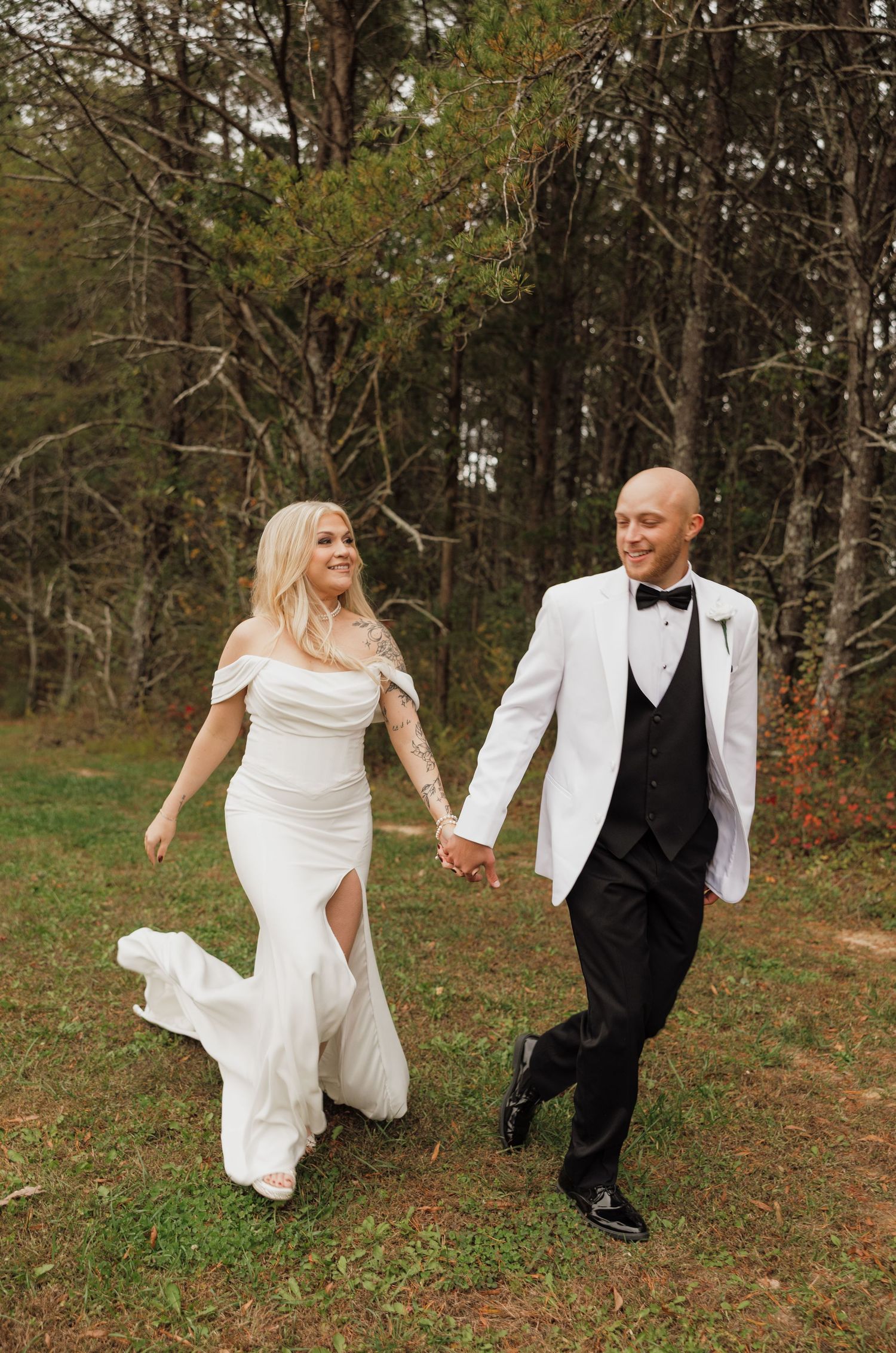 A couple in wedding attire playfully kicks up their feet while holding hands in a wooded outdoor setting.