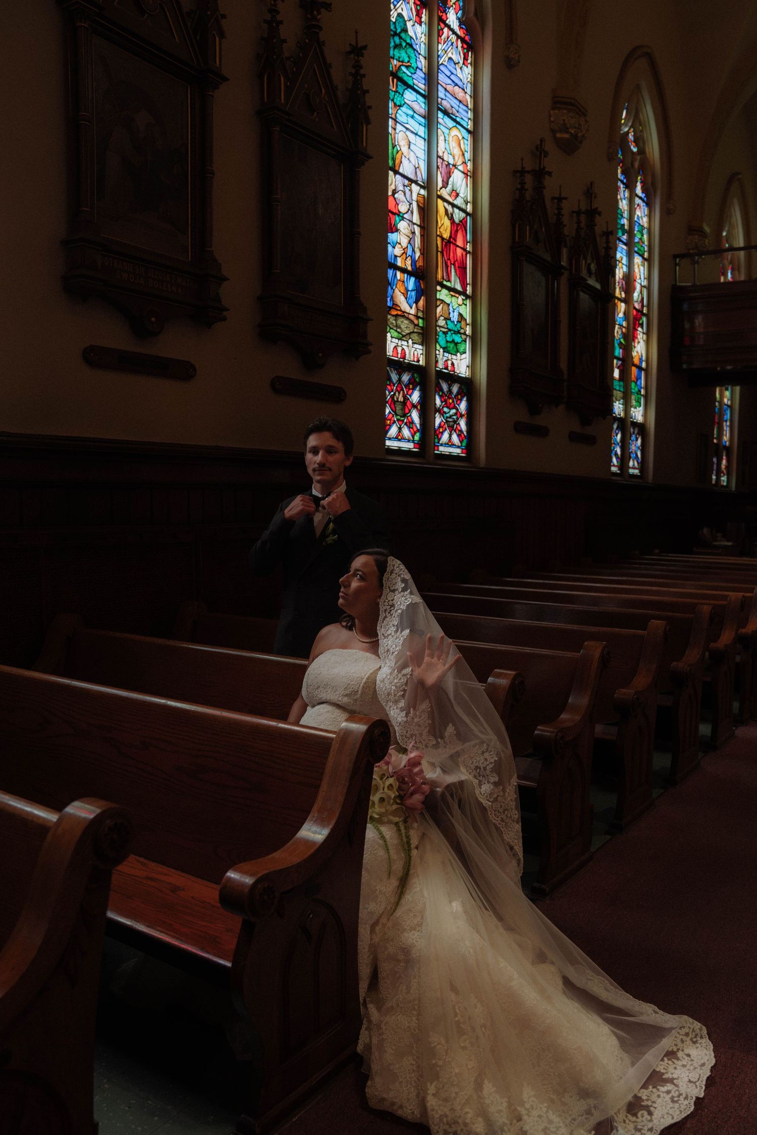 Bride and groom share a quiet moment in a dimly lit church with colorful stained glass windows above.
