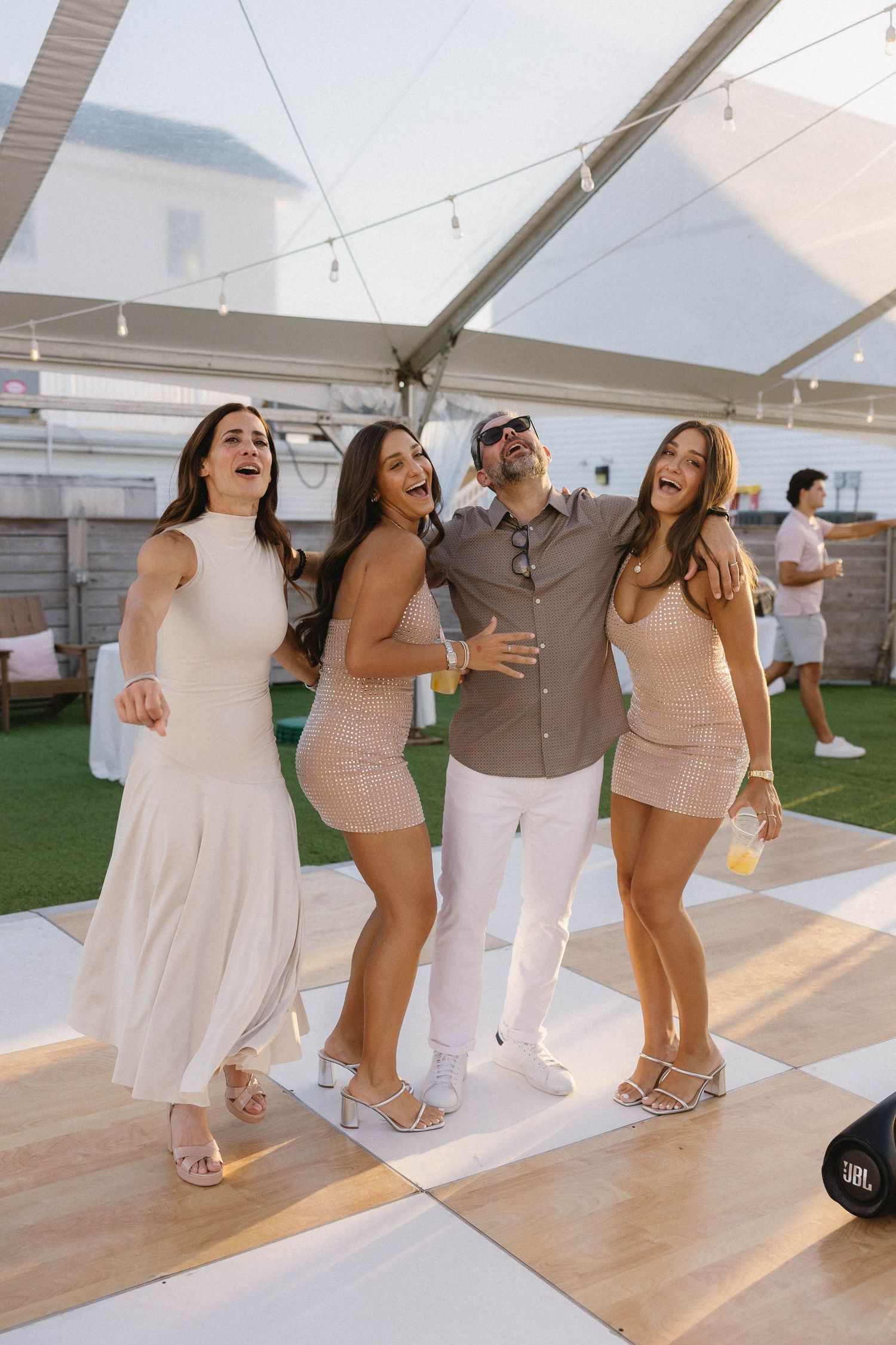 Group of four friends laughing and posing together on a checkered dance floor at an outdoor party.