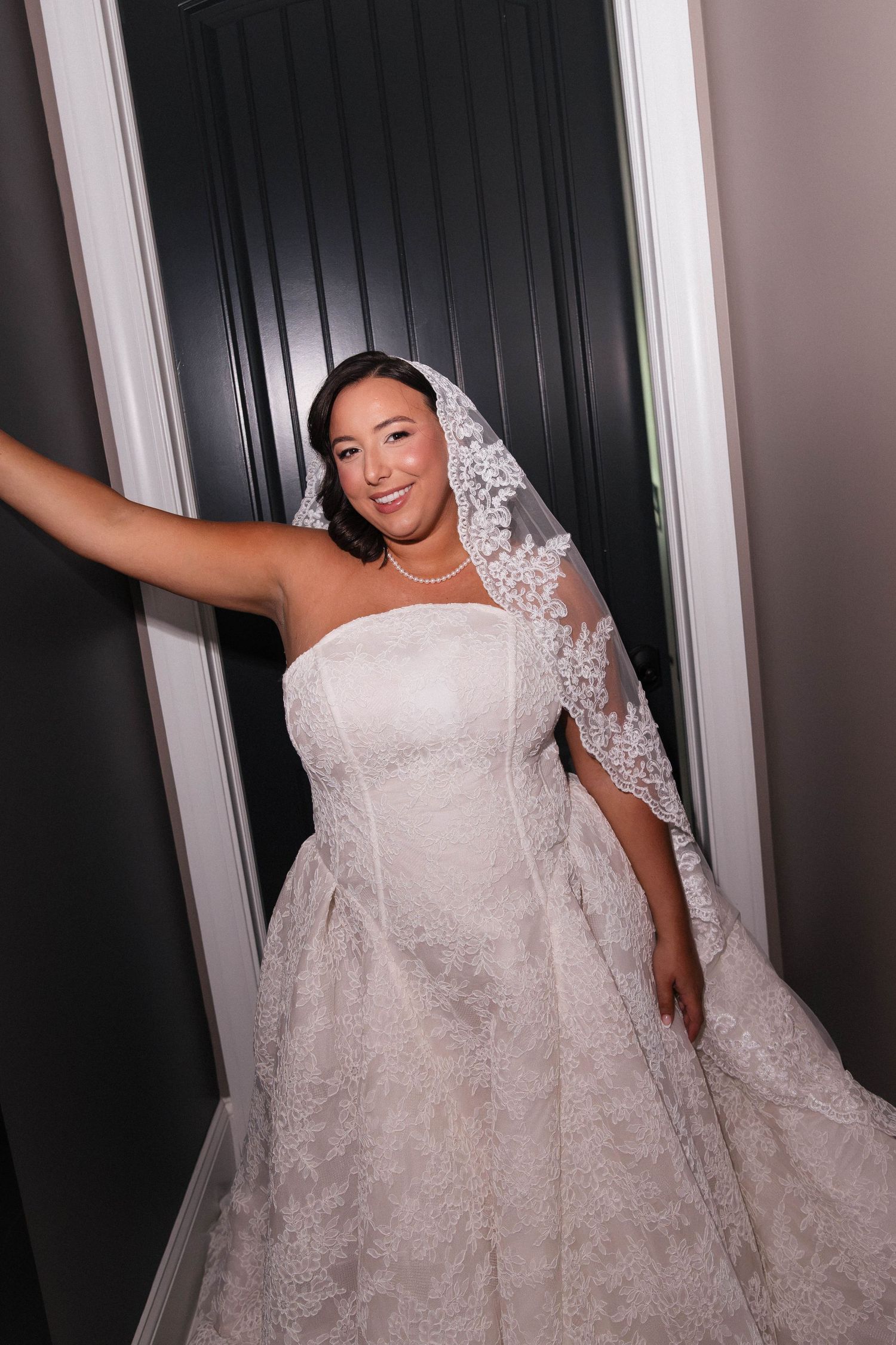 Smiling bride in strapless lace wedding gown and veil poses in a doorway.