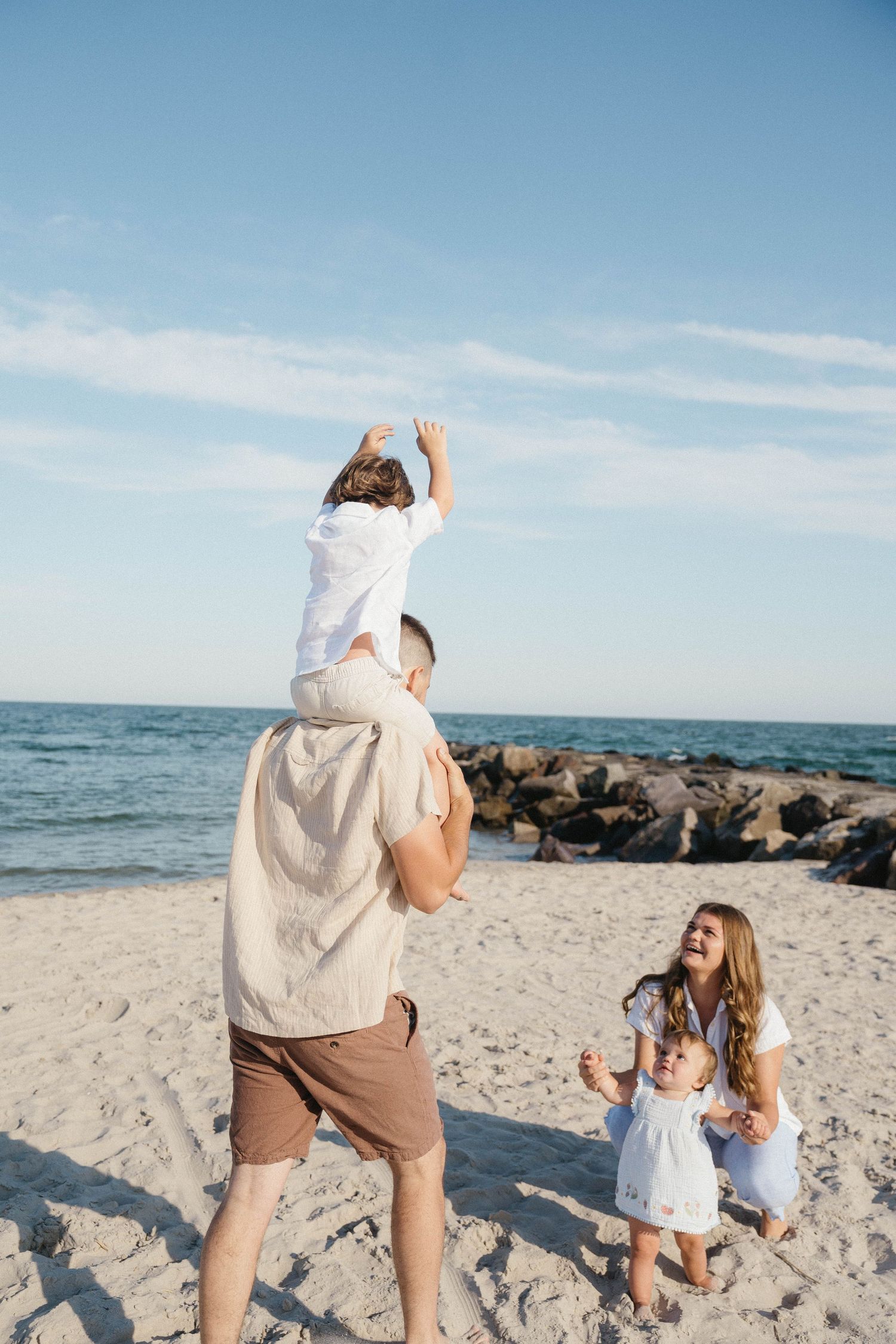 A joyful family enjoys a sunny beach day, with dad carrying a child on his shoulders while mom holds a baby nearby.