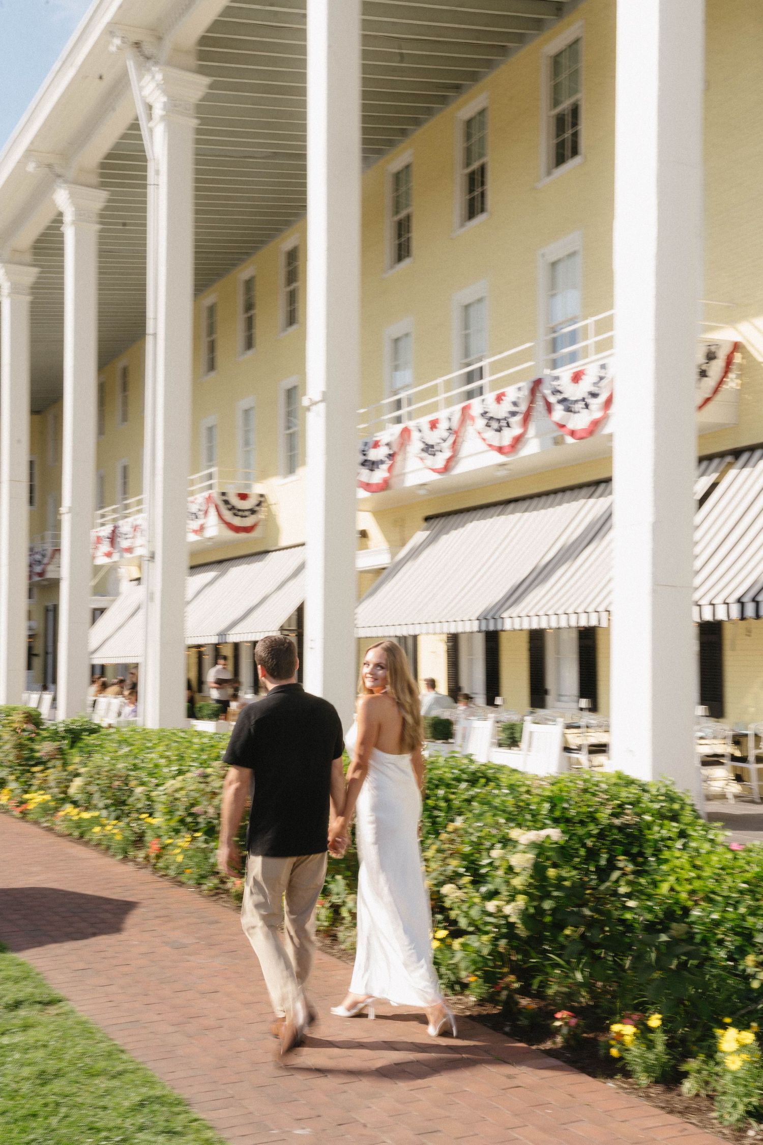 Couple walking hand in hand past a grand yellow colonial hotel with patriotic bunting decorations.