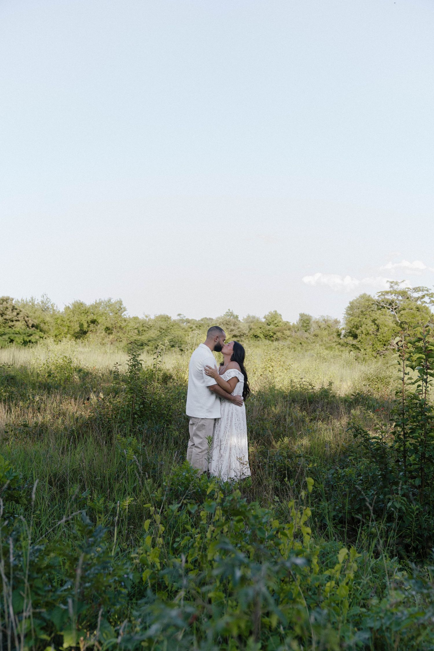 Couple embracing in a lush green field, wearing white outfits, sharing a romantic moment outdoors.
