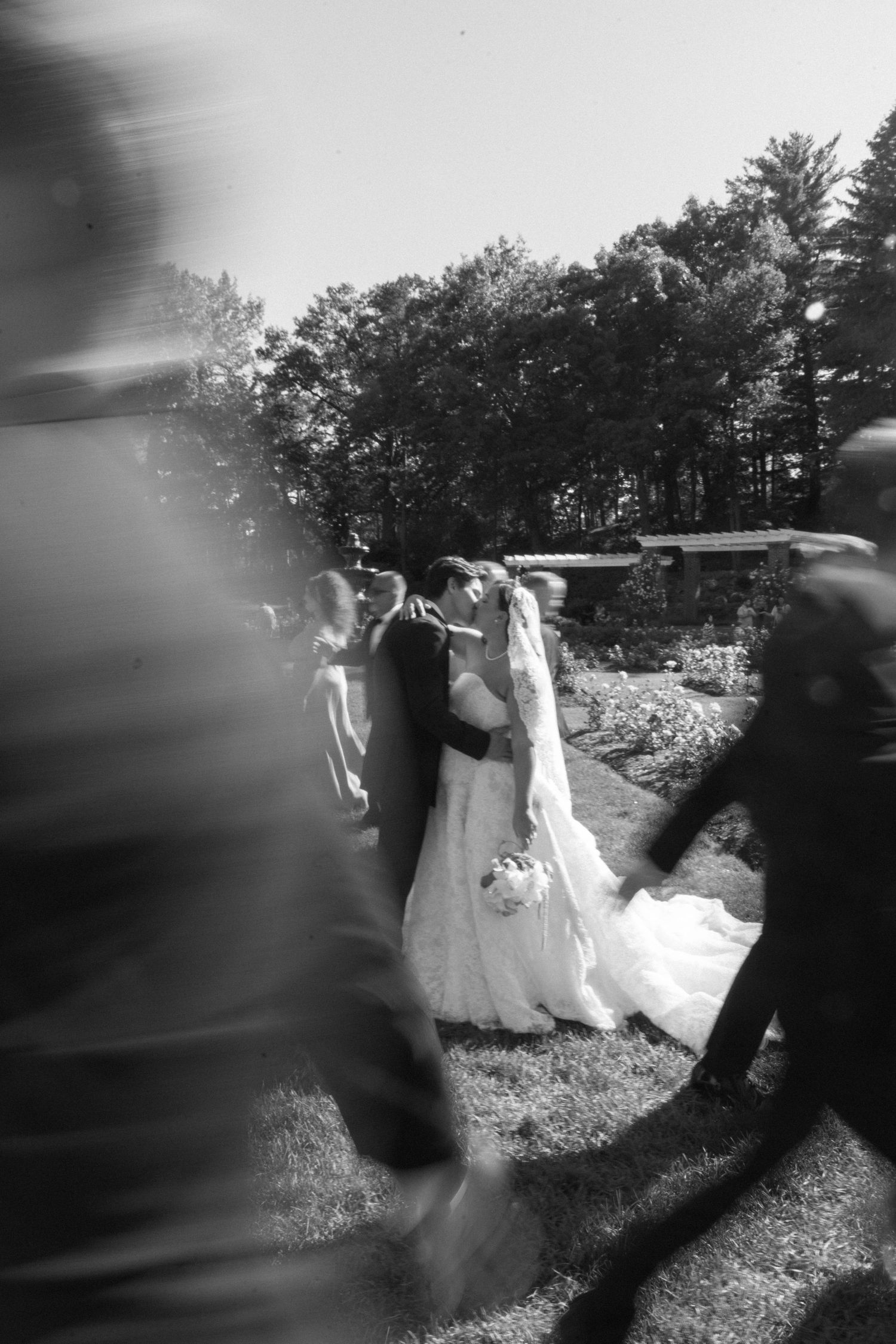 Black and white candid shot of newlyweds kissing outdoors as wedding guests move around them in blur.