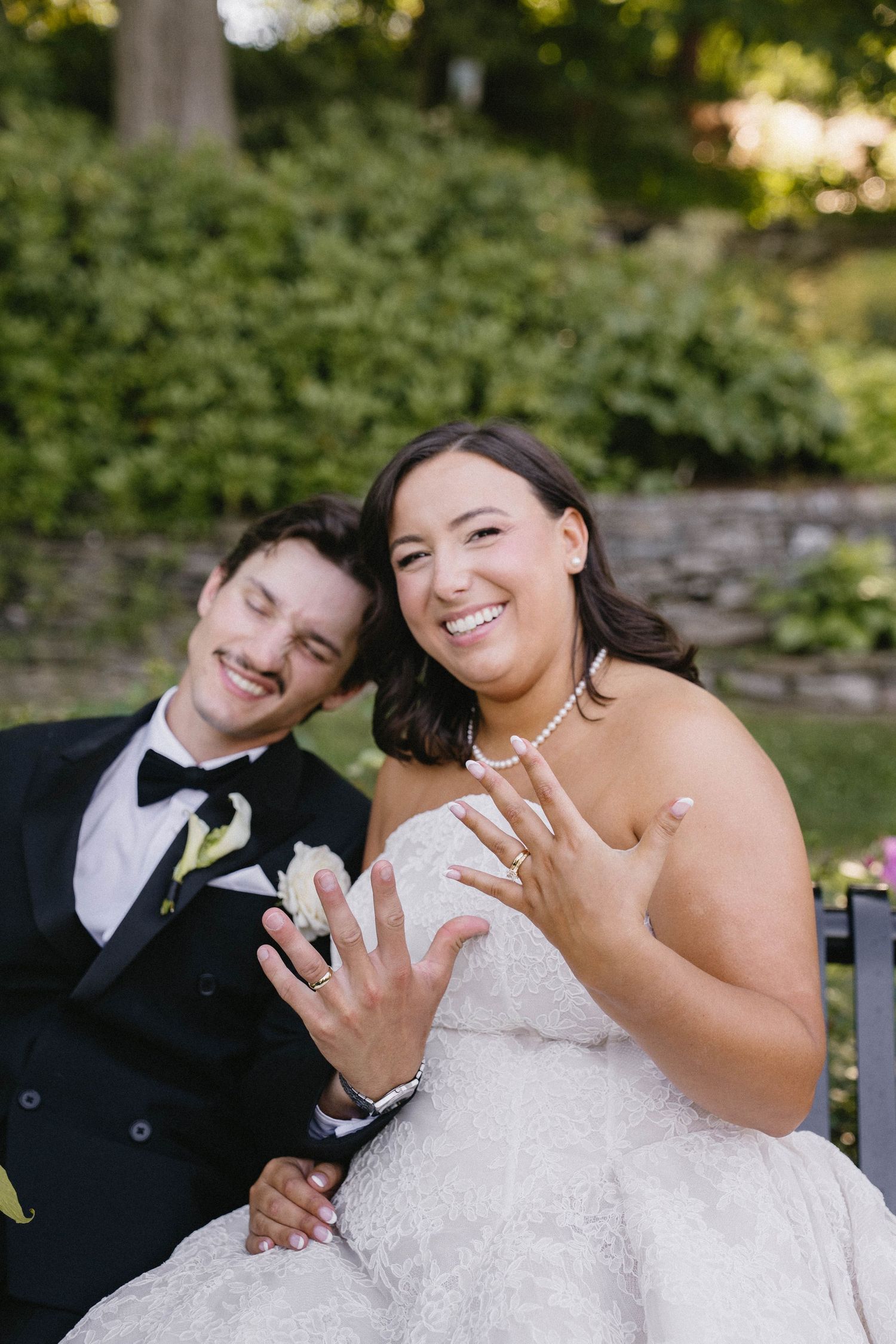 Newlyweds smiling and showing off wedding rings while sitting outdoors, bride in strapless gown and groom in black tuxedo.