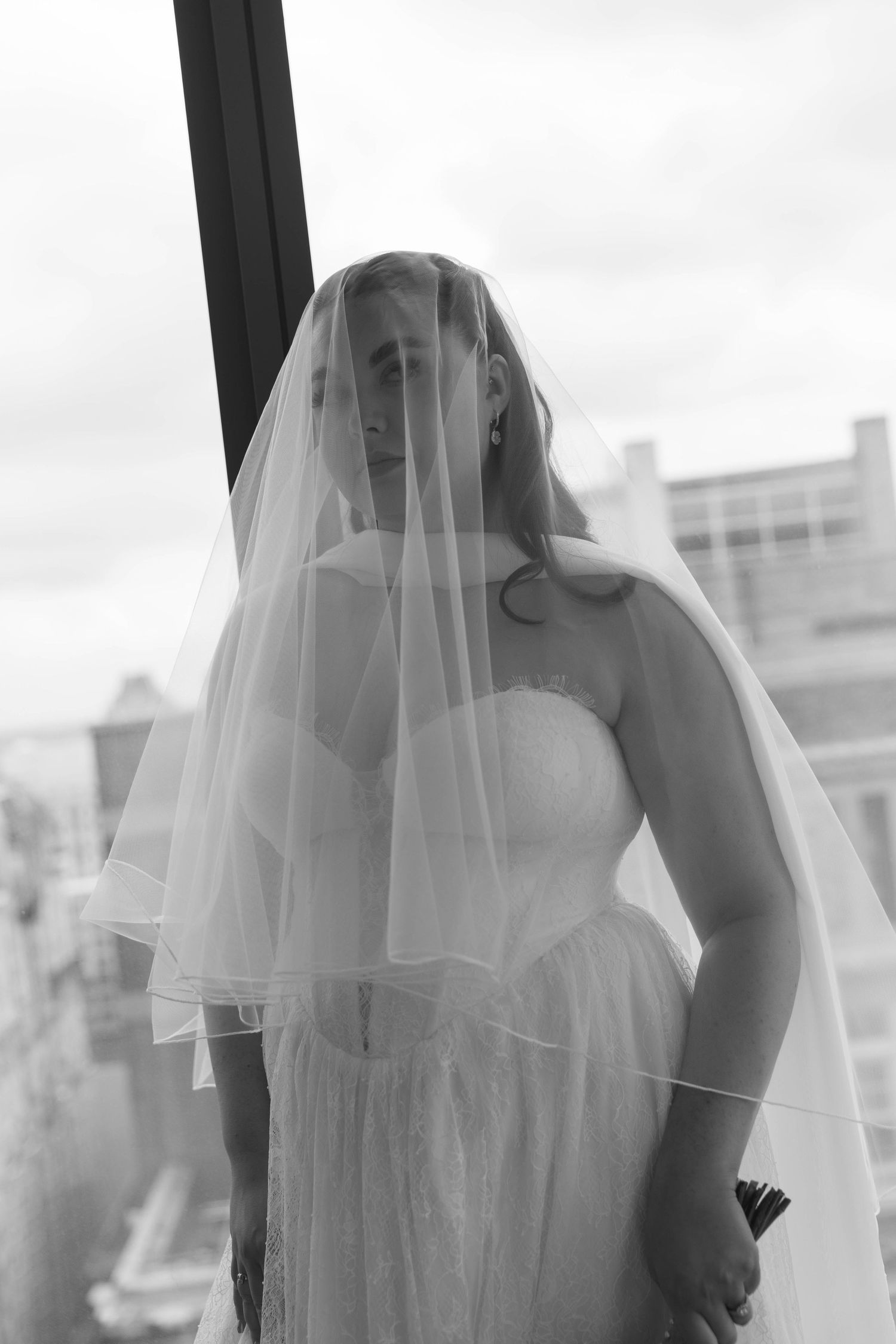 Black and white portrait of a bride wearing a flowing veil and strapless gown, standing near a window with city views.
