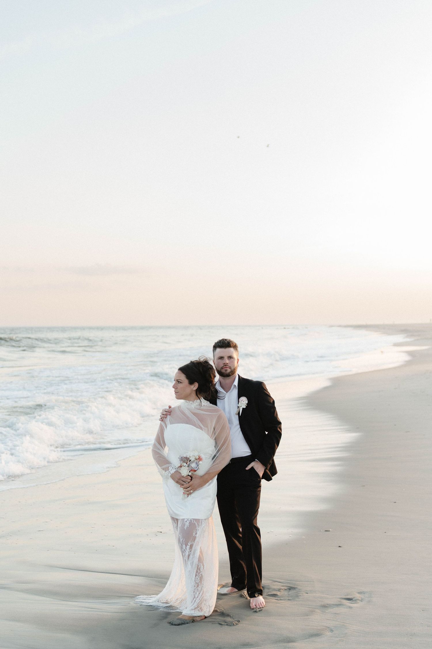 Bride and groom embrace on a sandy beach at sunset, waves crashing behind them, she holds a bouquet.