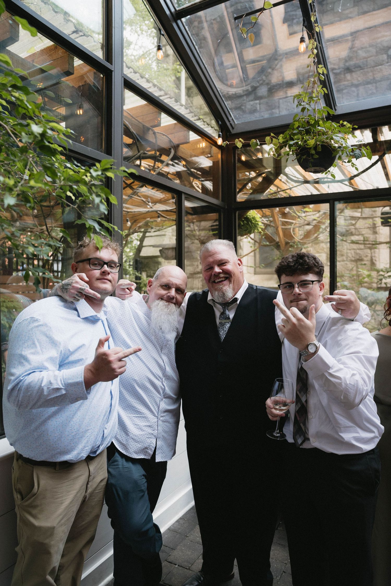 Four men posing together at an indoor wedding venue with glass ceiling and greenery, making hand gestures.