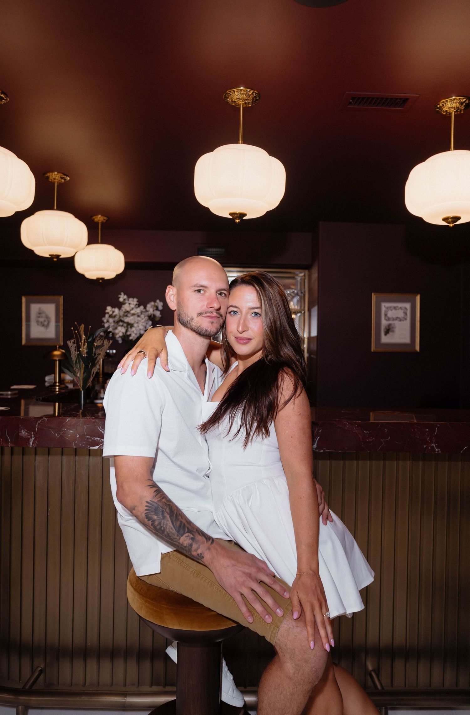 A couple in matching white outfits poses intimately at a bar with warm globe pendant lights.