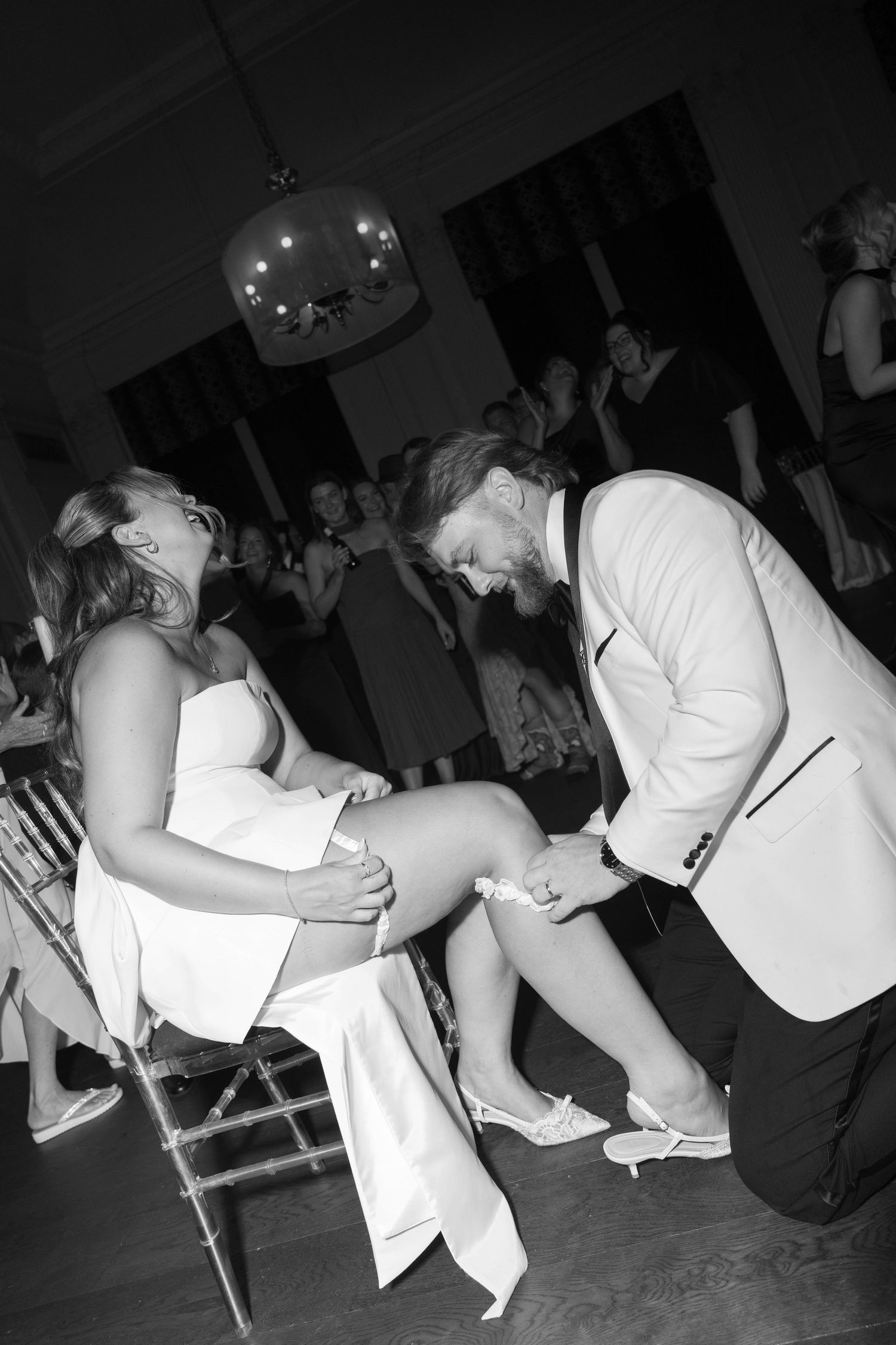 Black and white photo of groom removing bride's garter at wedding reception while guests celebrate in background.
