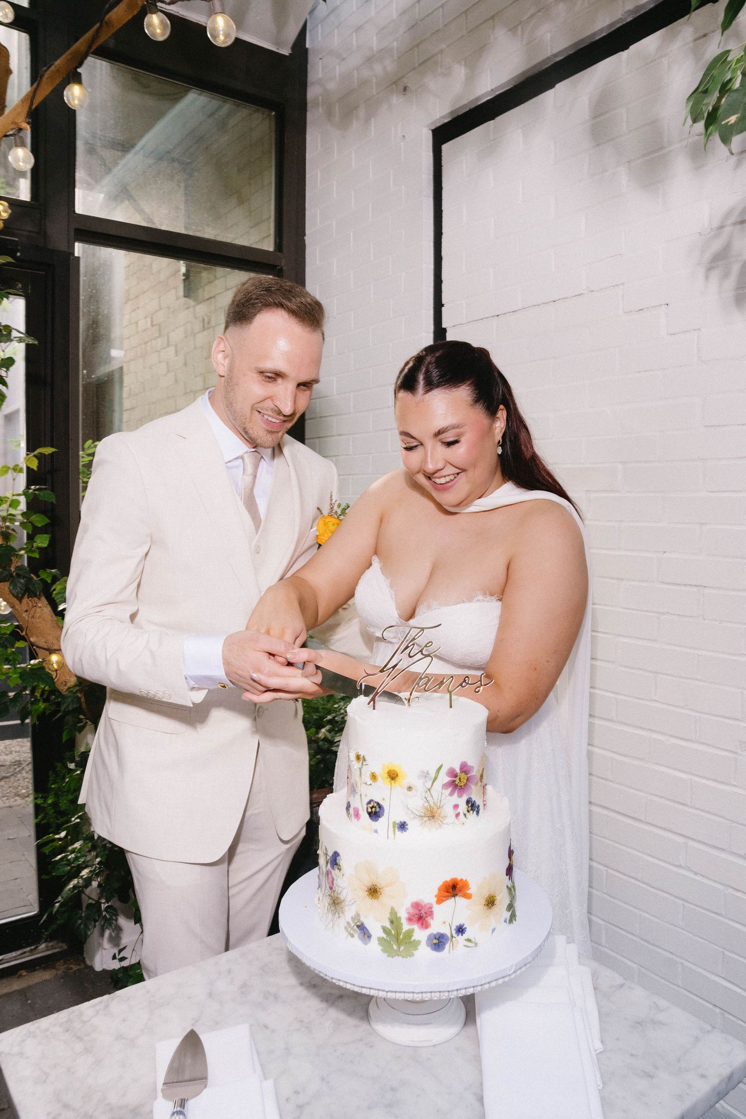 Bride and groom cutting a three-tier white wedding cake decorated with pressed flowers at their wedding reception.