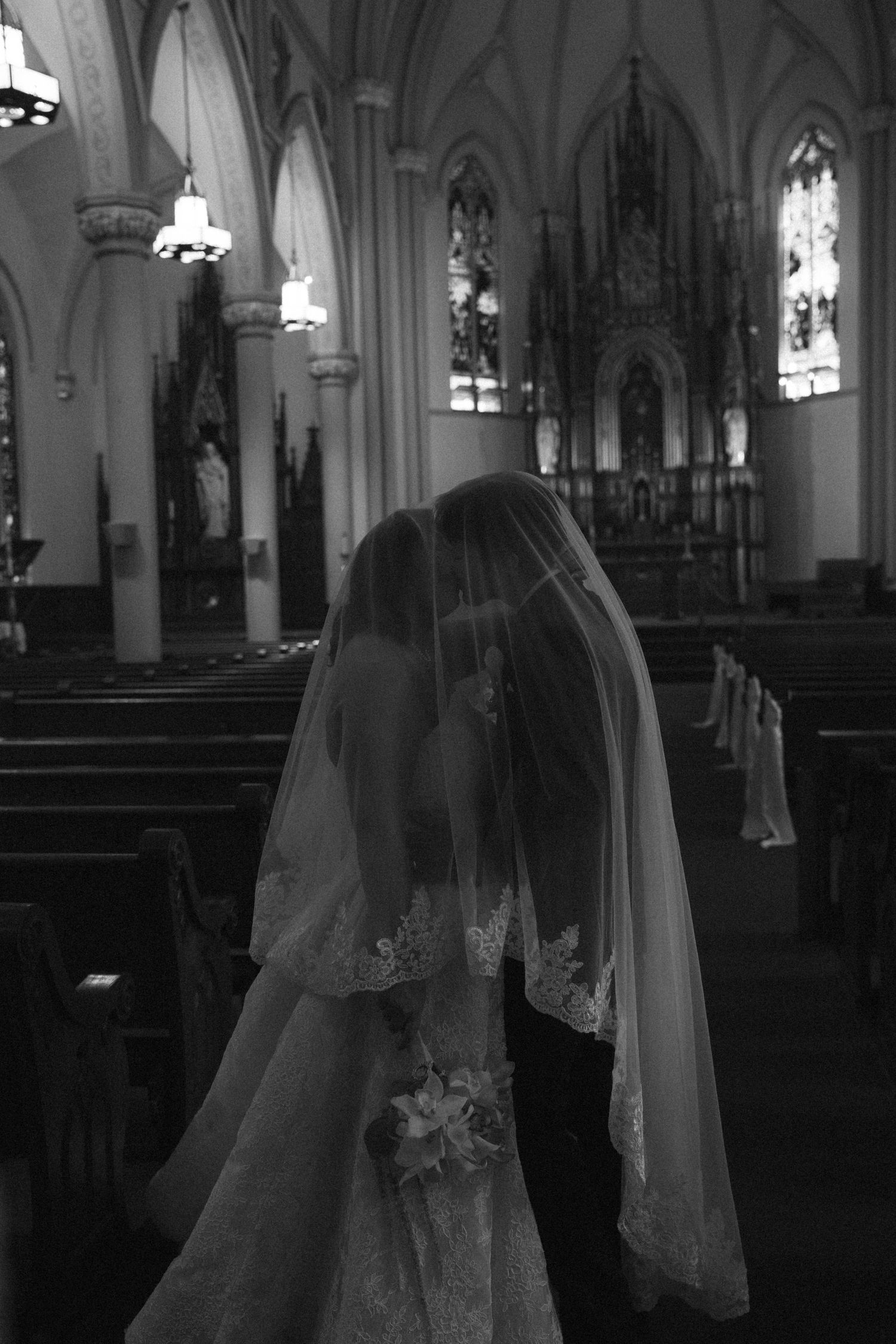 Black and white photo of bride and groom walking down aisle of ornate church under veil.