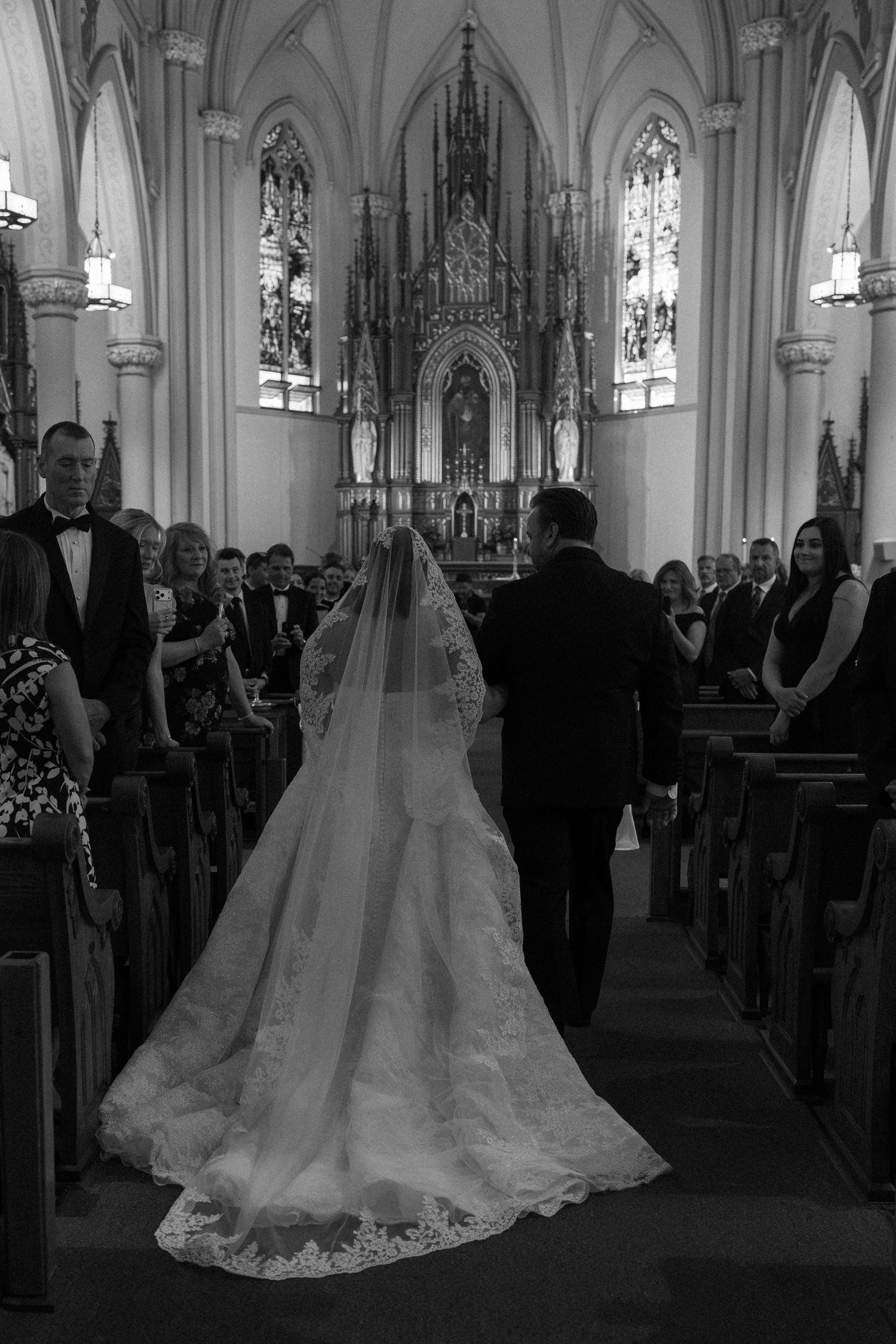 Bride walking down church aisle in elegant lace gown and cathedral veil, black and white wedding ceremony photo.
