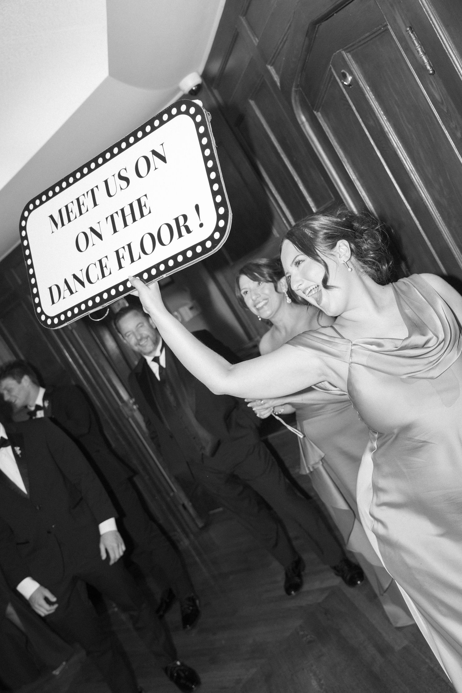 Bridesmaid holding Meet Us On The Dance Floor sign with wedding party in black and white photo.