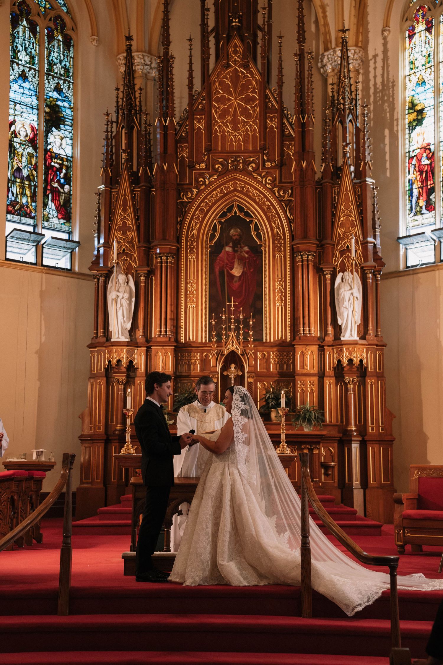 A bride and groom exchange vows at an ornate church altar with stained glass windows and golden Gothic woodwork.