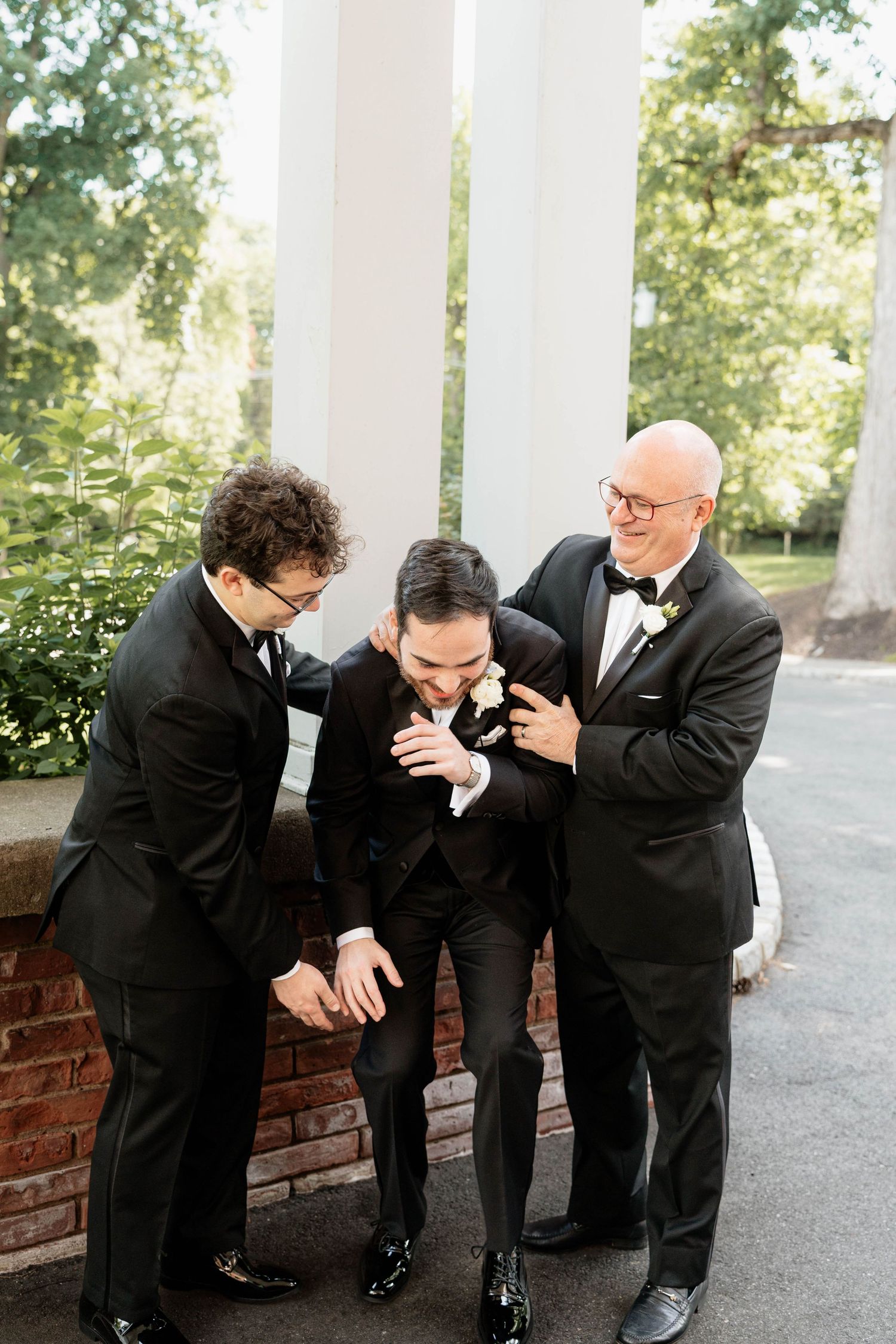Three groomsmen in black tuxedos share a joyful laugh together outside near white columns.