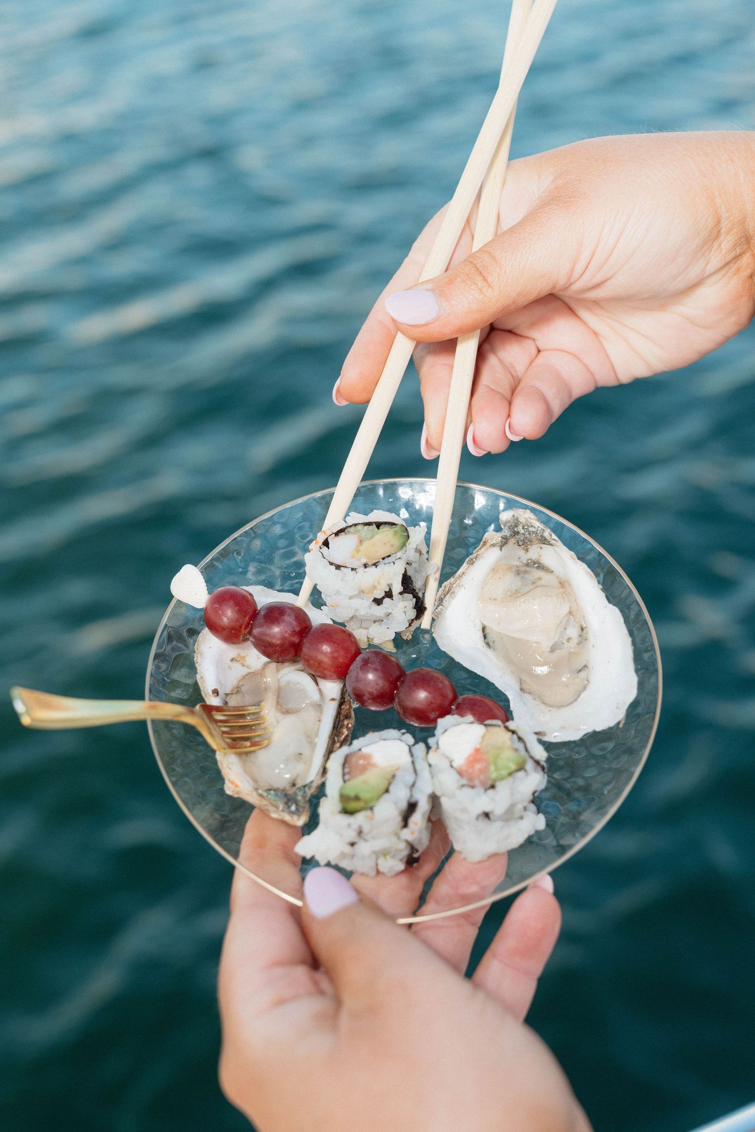 Hands holding glass plate of sushi rolls and oysters with chopsticks over ocean water.