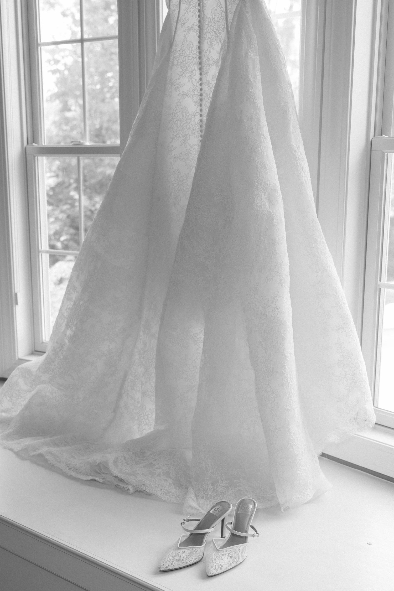 Black and white photo of a lace wedding dress hanging by a window with bridal heels on the sill below.