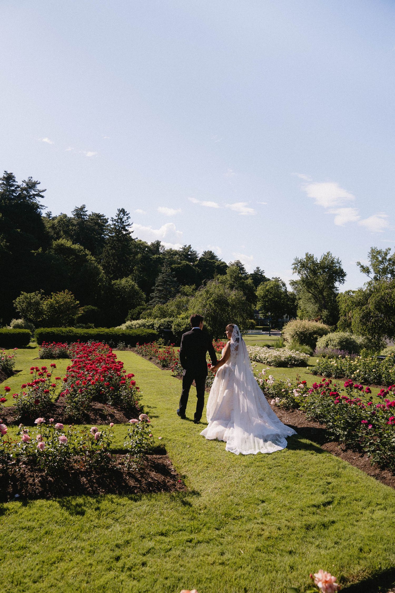 Bride and groom walking hand in hand through a lush rose garden on their wedding day.