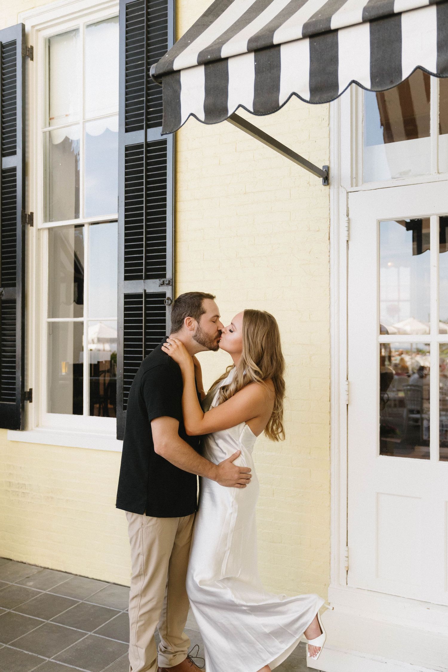 Couple sharing a romantic kiss outside a yellow brick building with black shutters and striped awning.