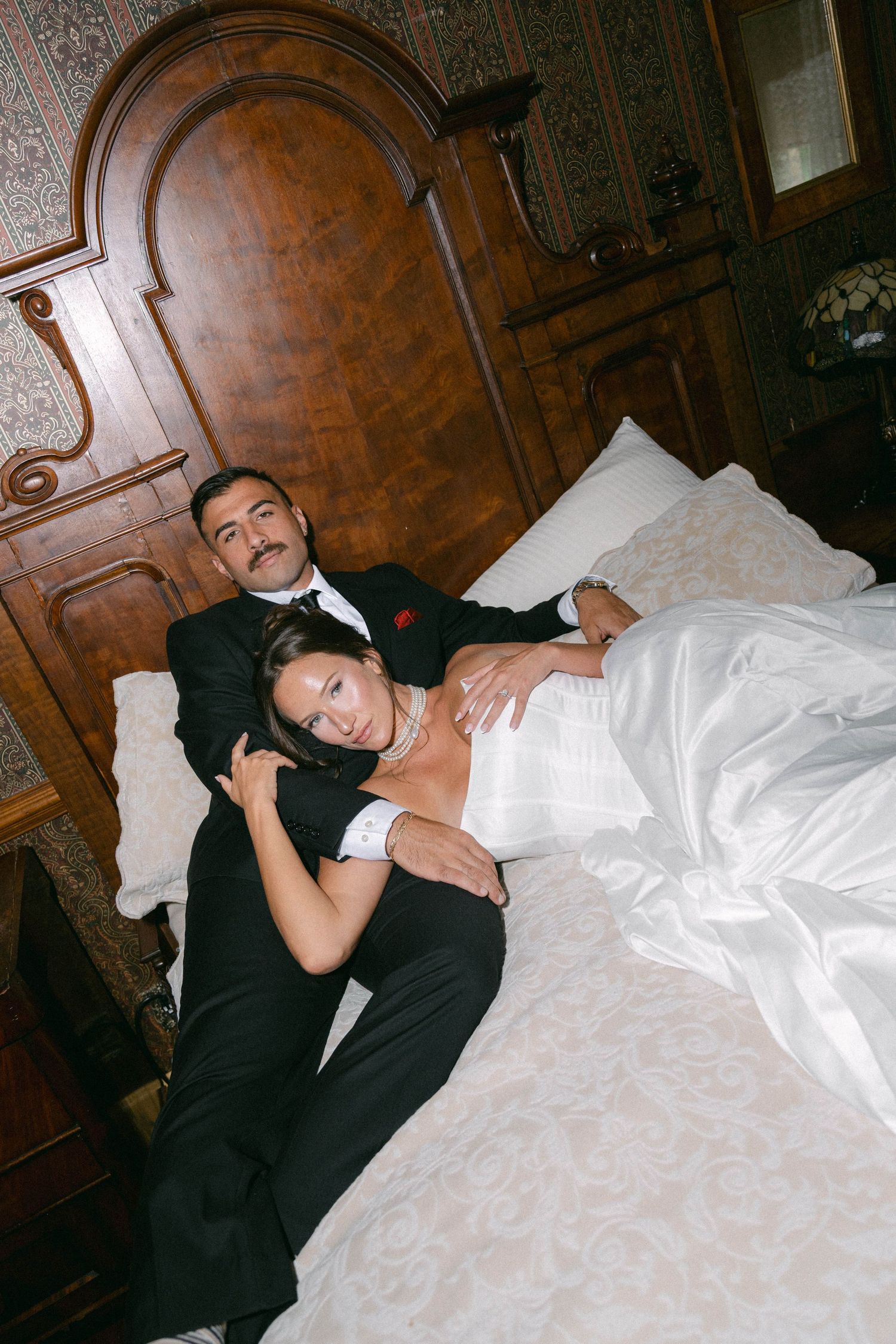 Bride and groom in wedding attire relaxing together on an elegant bed with ornate wooden headboard.