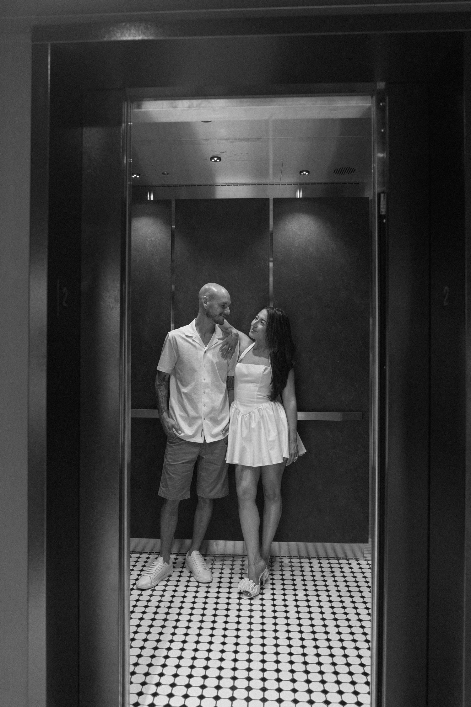 Black and white photo of a couple sharing a tender moment inside an elevator with checkered tile flooring.