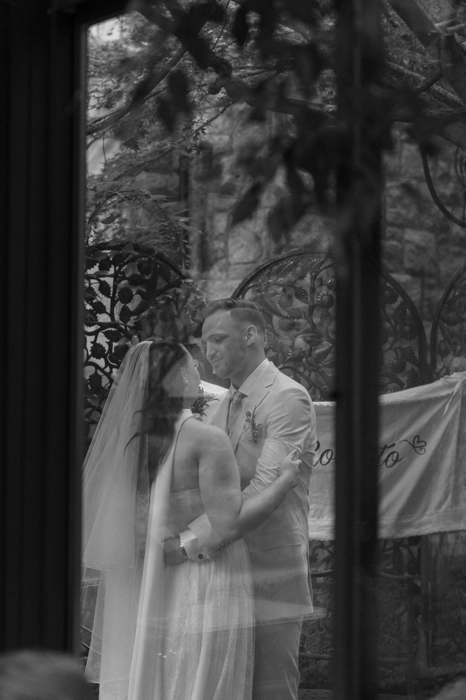 Black and white wedding photo of bride and groom sharing intimate moment, viewed through glass window.